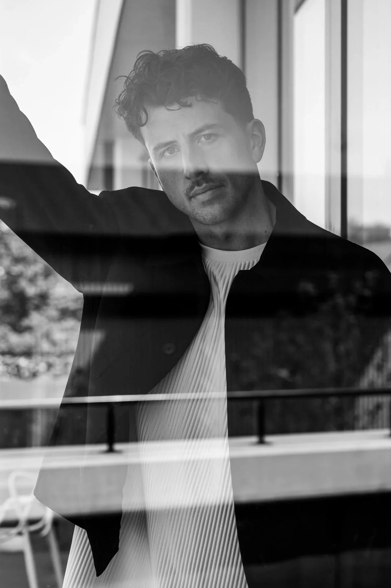 Een man met een krullende haardos en een baardje en snor, die door een raam kijkt. De foto is zwartwit en er is een reflectie van de omgeving op het raam.