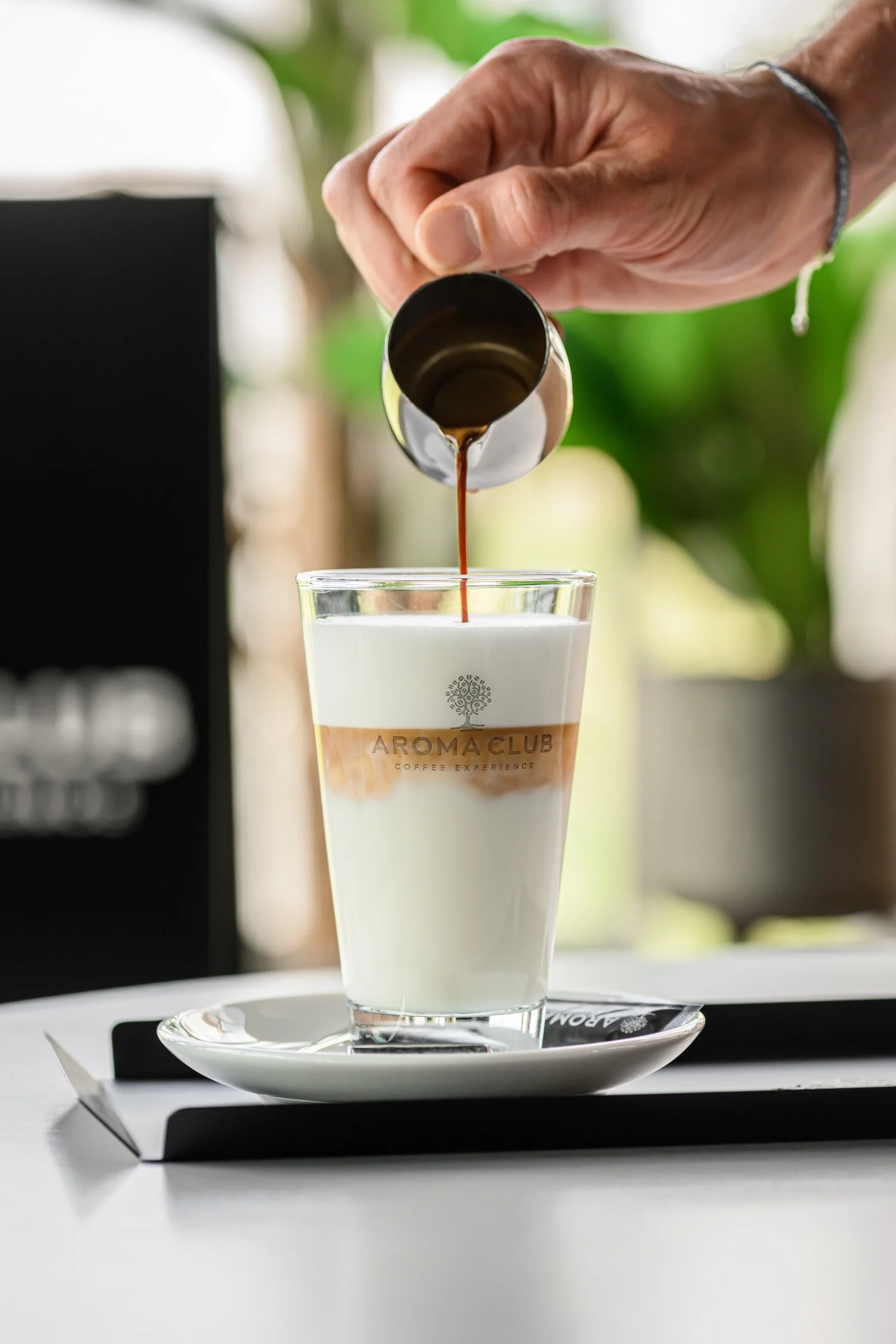 Persoon giet koffie in een glas met melk, met 'AROMA CLUB' op het glas, op een dienblad op een witte tafel.