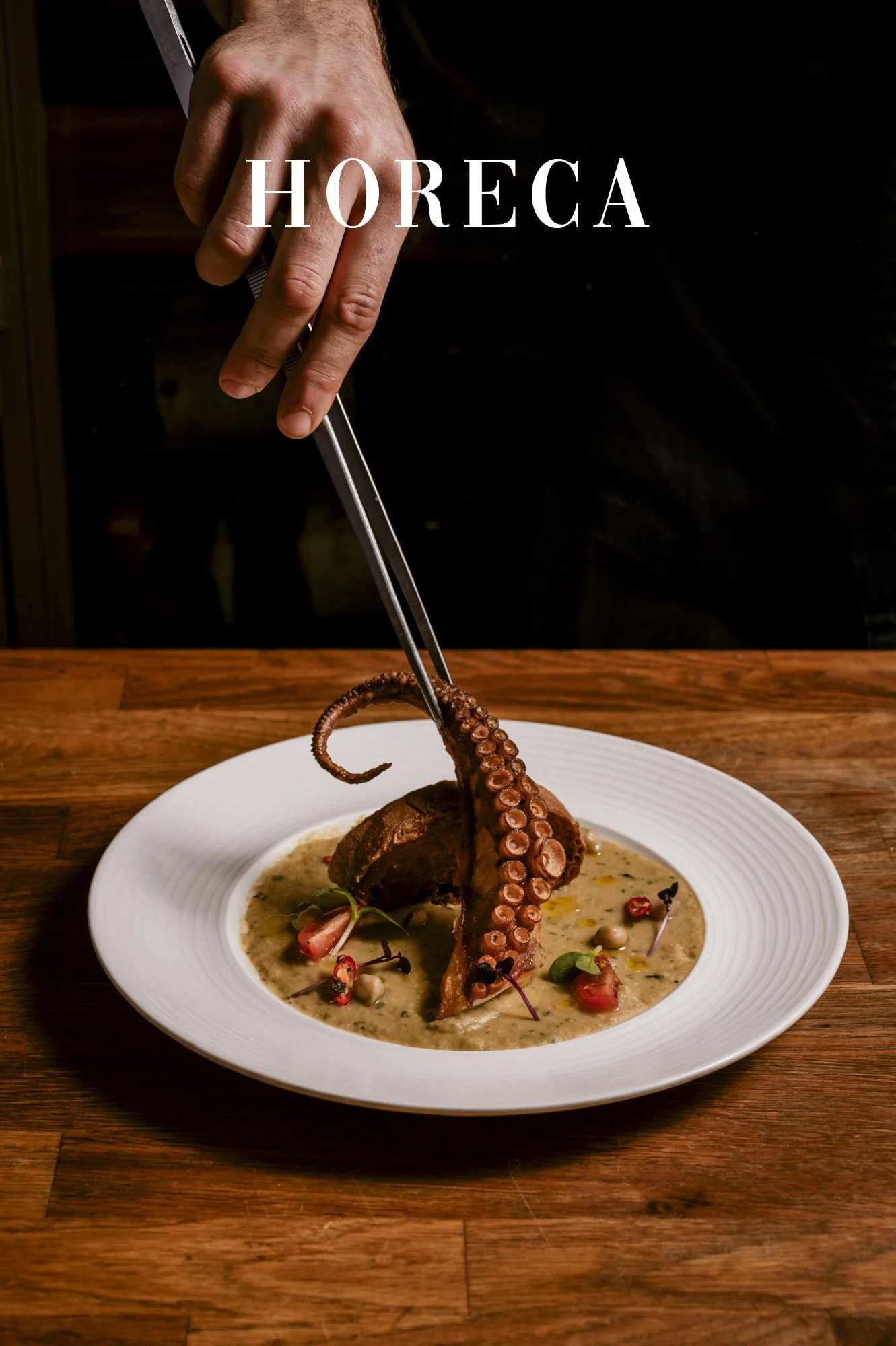 Een ober gebruikt tangen om een gegrilde octopus op een bord te plaatsen, dat in een warme roomsaus ligt, versierd met kleine bloemen en kruiden.