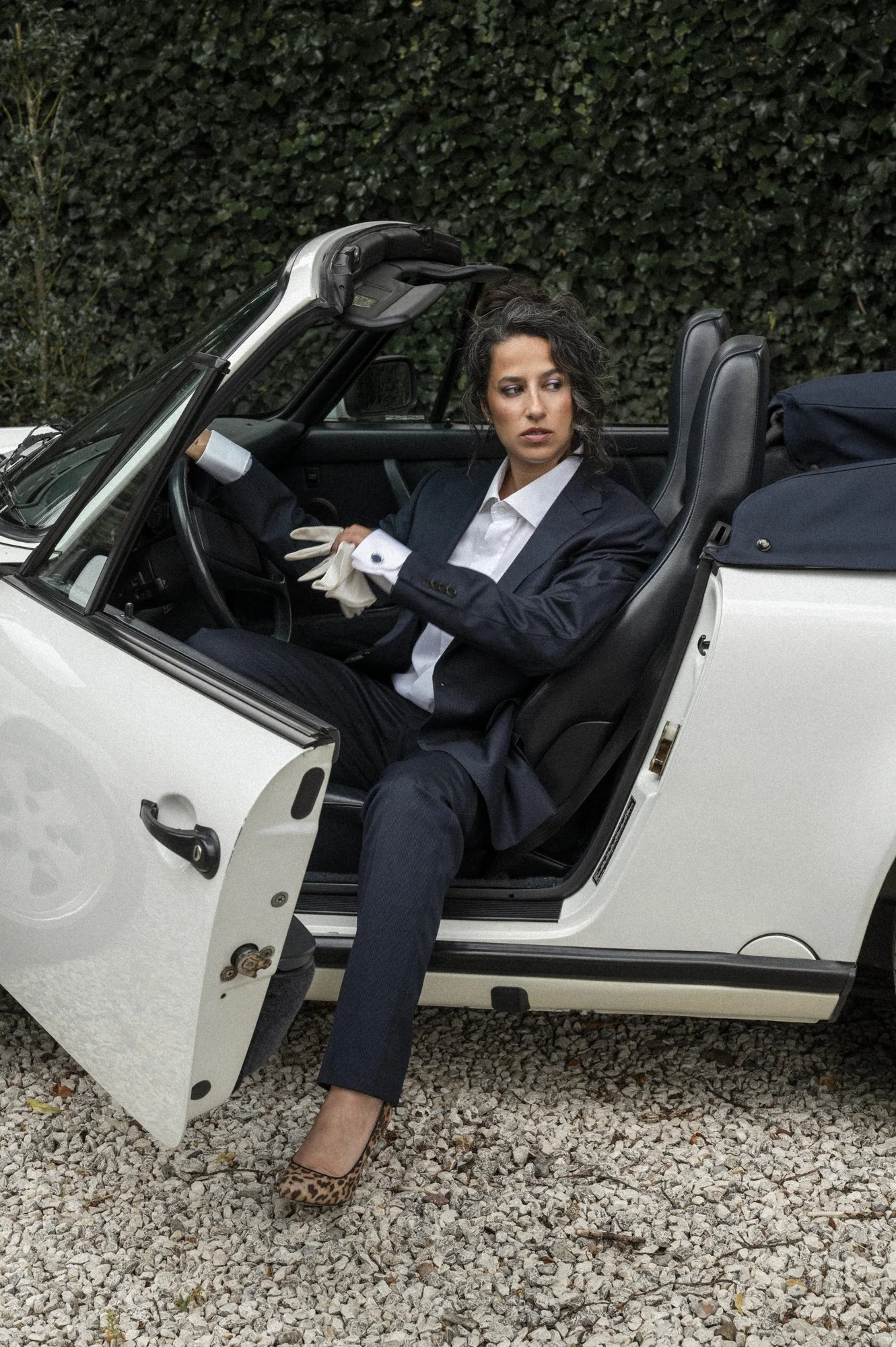 Vrouw in een zwart pak en witte blouse zit in een witte cabriolet auto, met de deur open en een bosje groen struik achter haar.