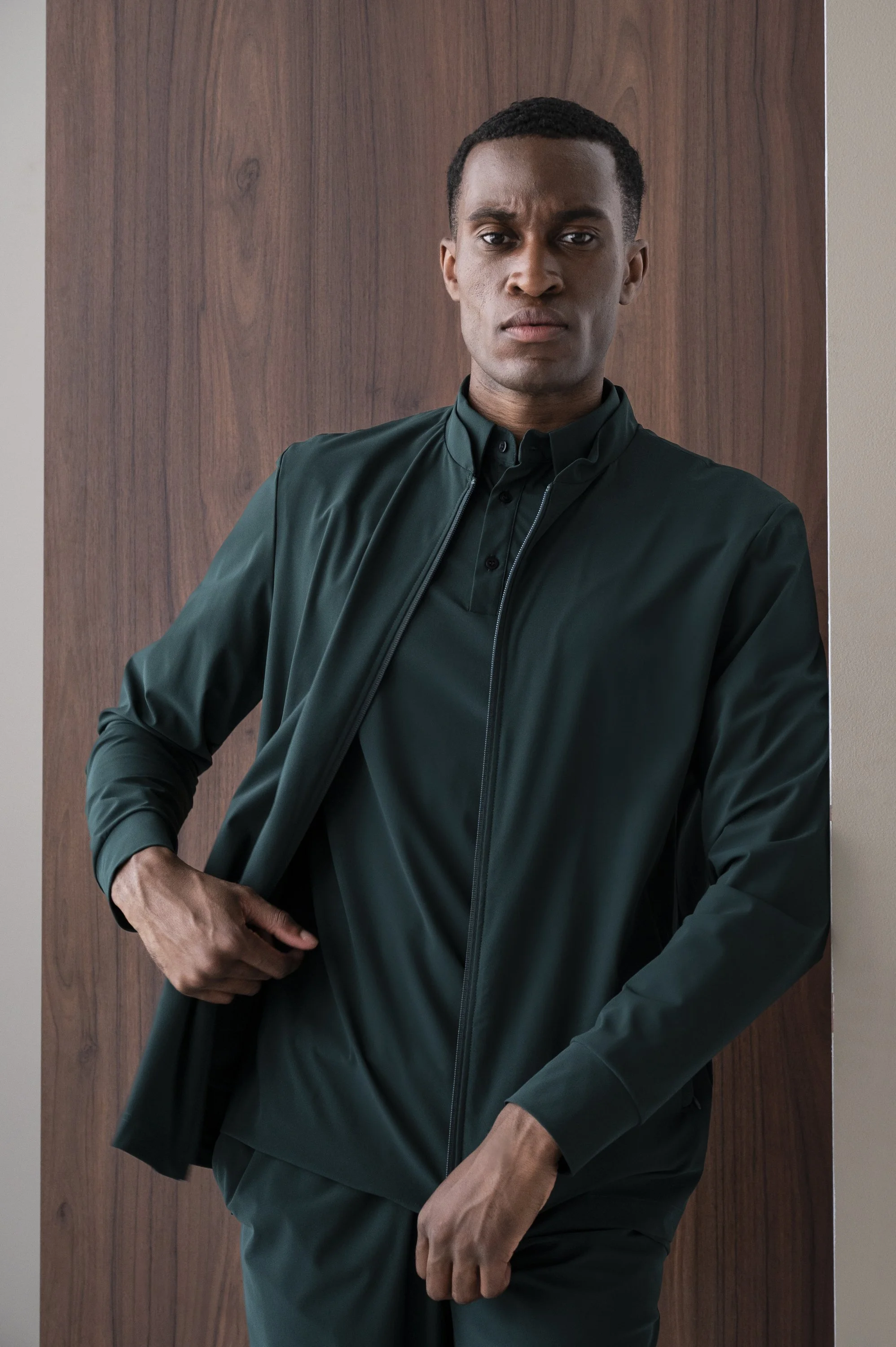 Man staat voor een houten muur, draagt een donkere groene jas en een zwarte shirt, heeft een serieuze uitdrukking en plaatst één hand in zijn taille.