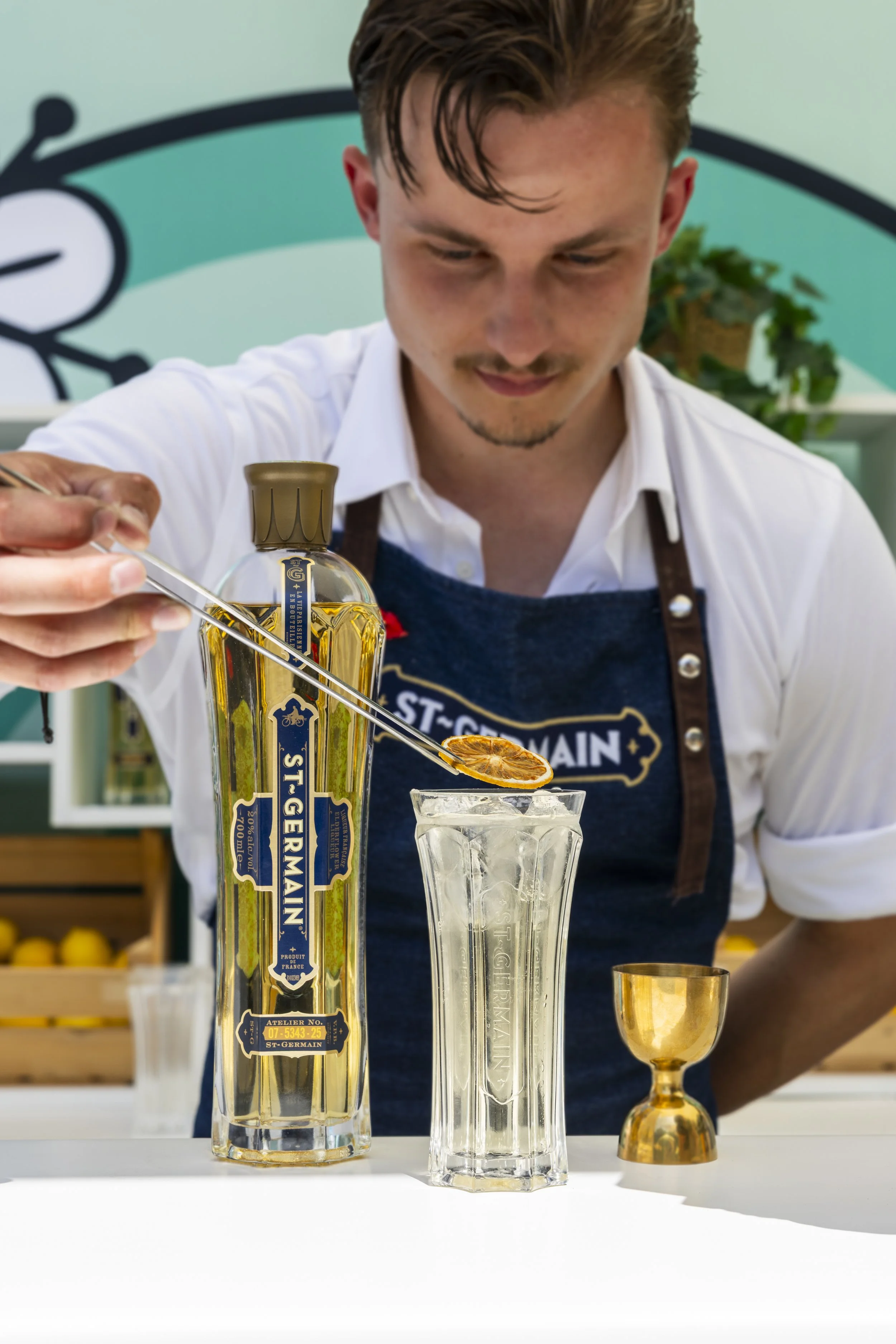 Een jonge barman legt een gedroogde citroen bovenop een fris ogende cocktail van St. Germain likeur, aan de bar in het zomerse zonlicht.