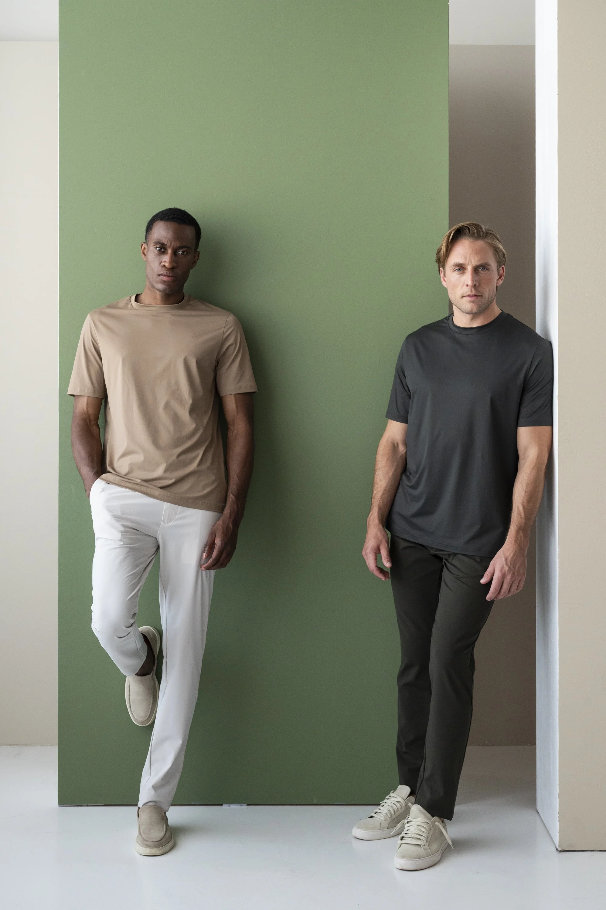 Twee mannelijke modellen staan tegen een groene en beige muur in een fotostudio, beiden gekleed in casual t-shirts en broek, één in beige en wit, de ander in zwart en zwart, en dragen lichte sneakers.