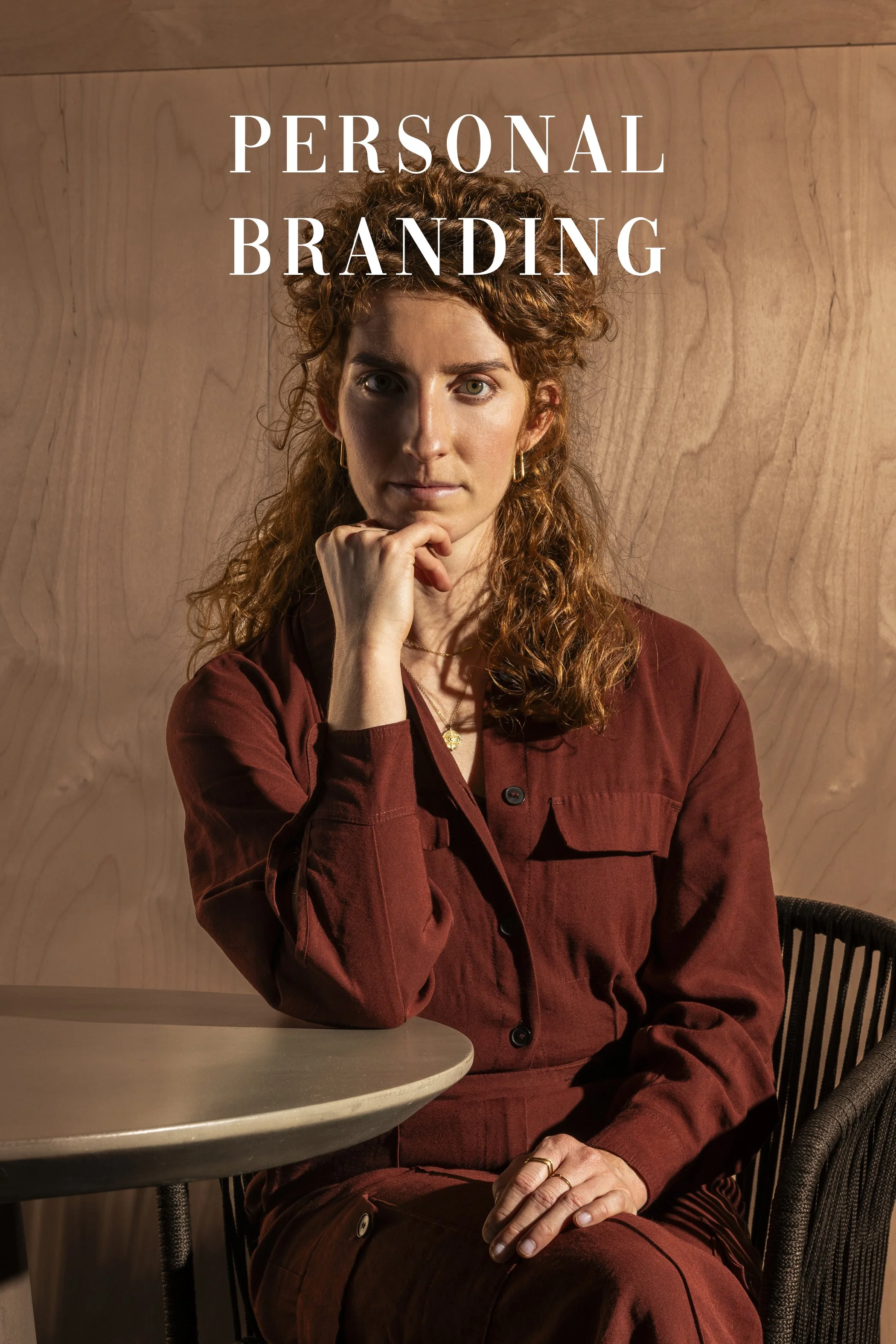 Portret van een vrouw met rood haar, zittend naast een ronde tafel, gekleed in een bruinachtig outfit, met een houten muur achter haar.