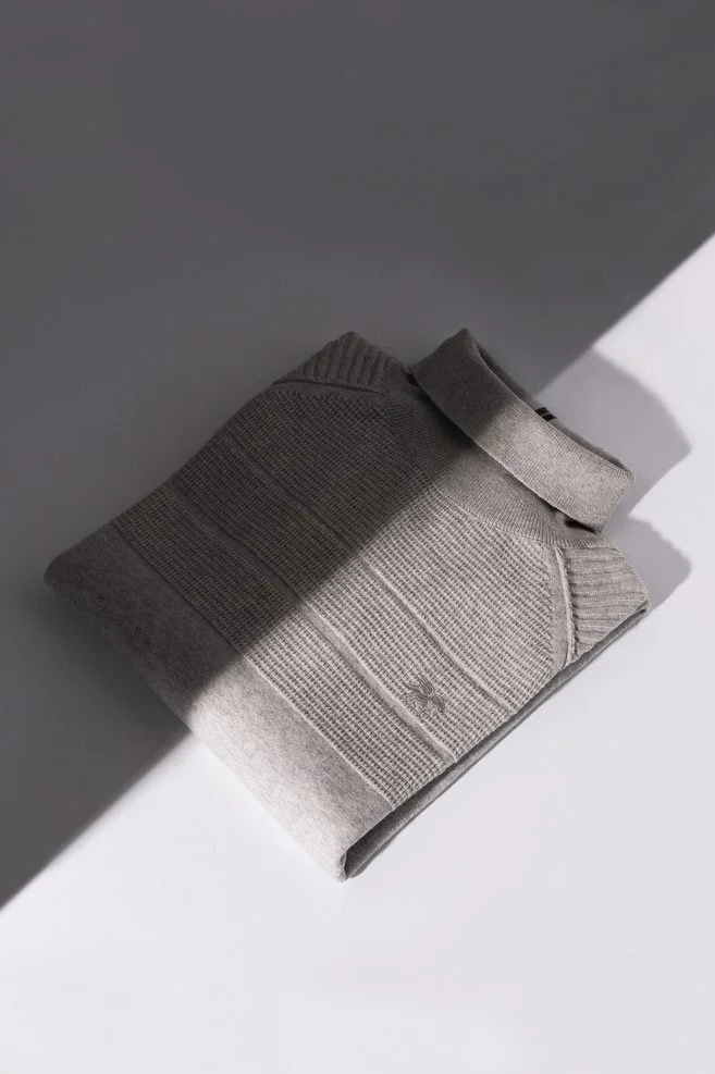 Grijze trui voor mannen, met geometrisch geweven patroon, op een witte ondergrond met een scherpe schaduw die het beeld splits in een donker en licht deel.