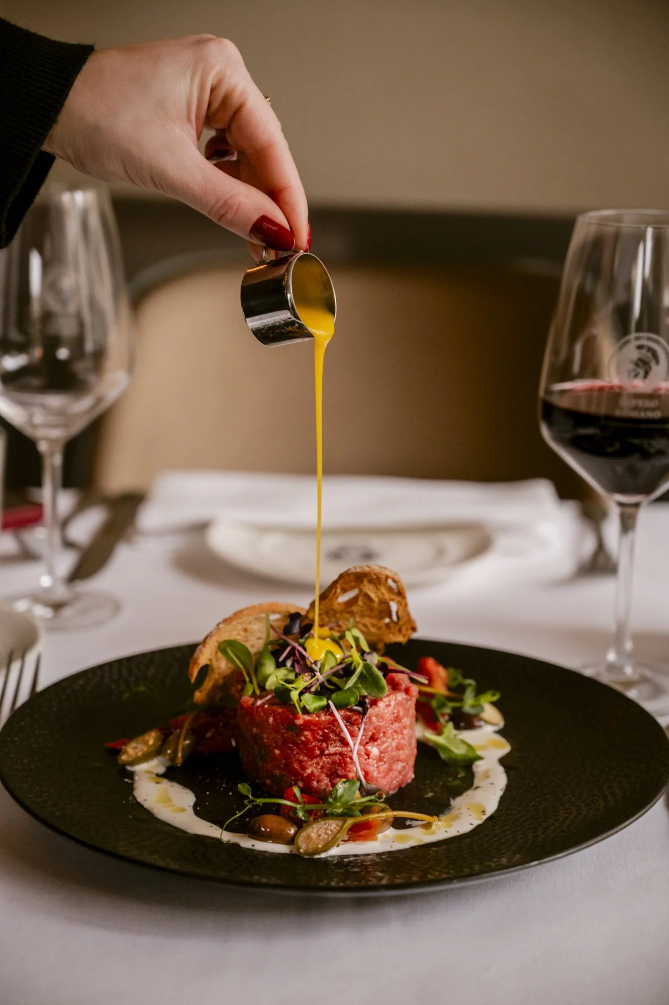 Hand giet eigeel op een gerecht met tartaar, groenten en microgreens, op een zwart stijlvol bord, omringd door wijnglazen op een gedekte tafel.