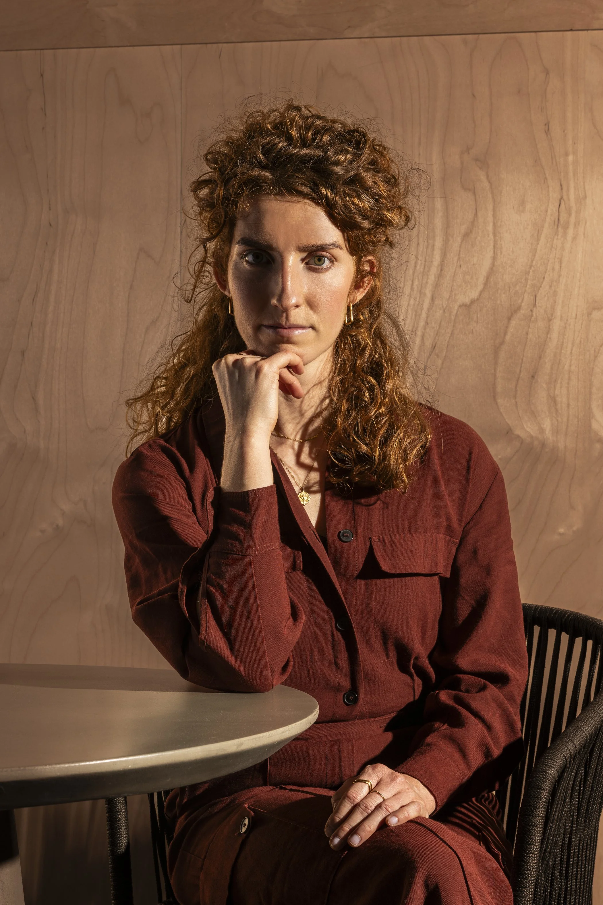 Portret van een vrouw met krullig, rood haar die in een interieur zit, leunt op haar hand en kijkt naar de camera.