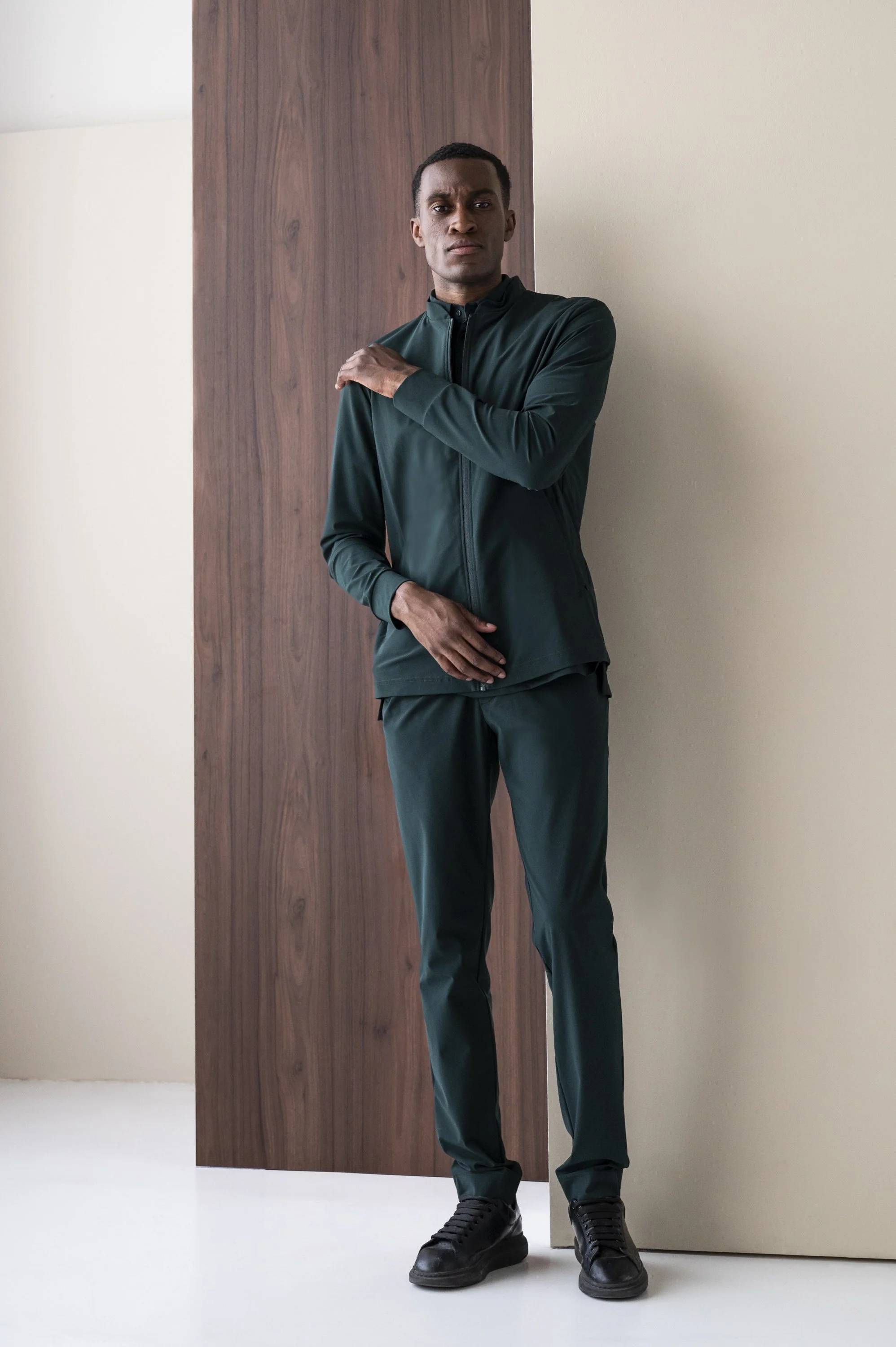 mannelijk model met donkere huidskleur poseert in donkergroene sportoutfit met schoenen en staat in een professionele fotostudio met houten en witte muur