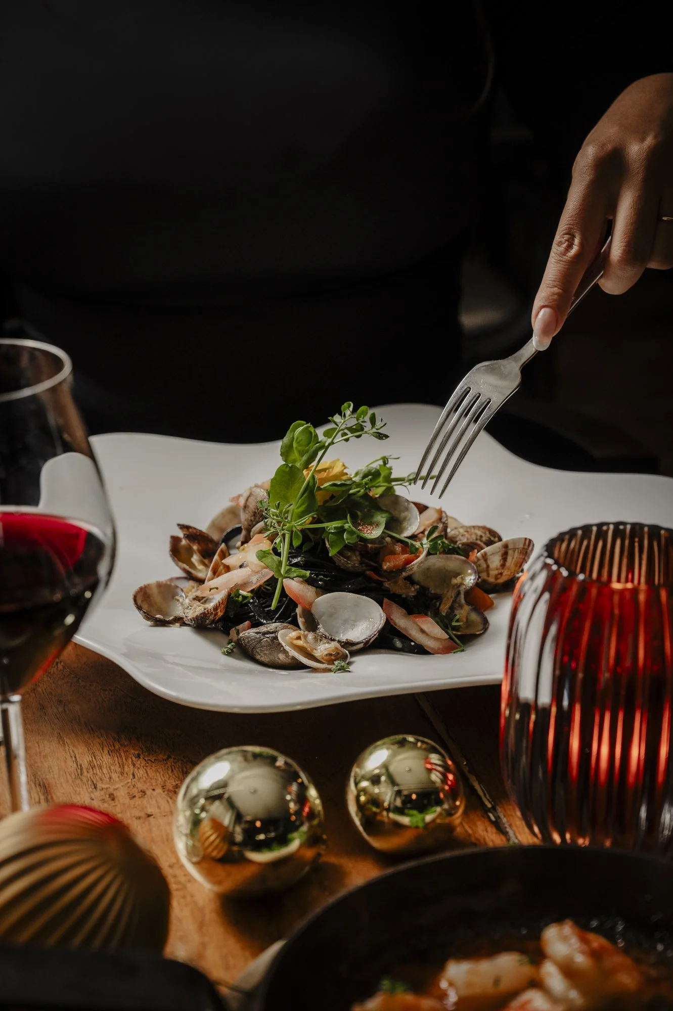 Een gerecht met zeevruchten op een wit bord, omringd door glazen wijn en decoratieve ballen. Een hand steekt met een vork in het gerecht.