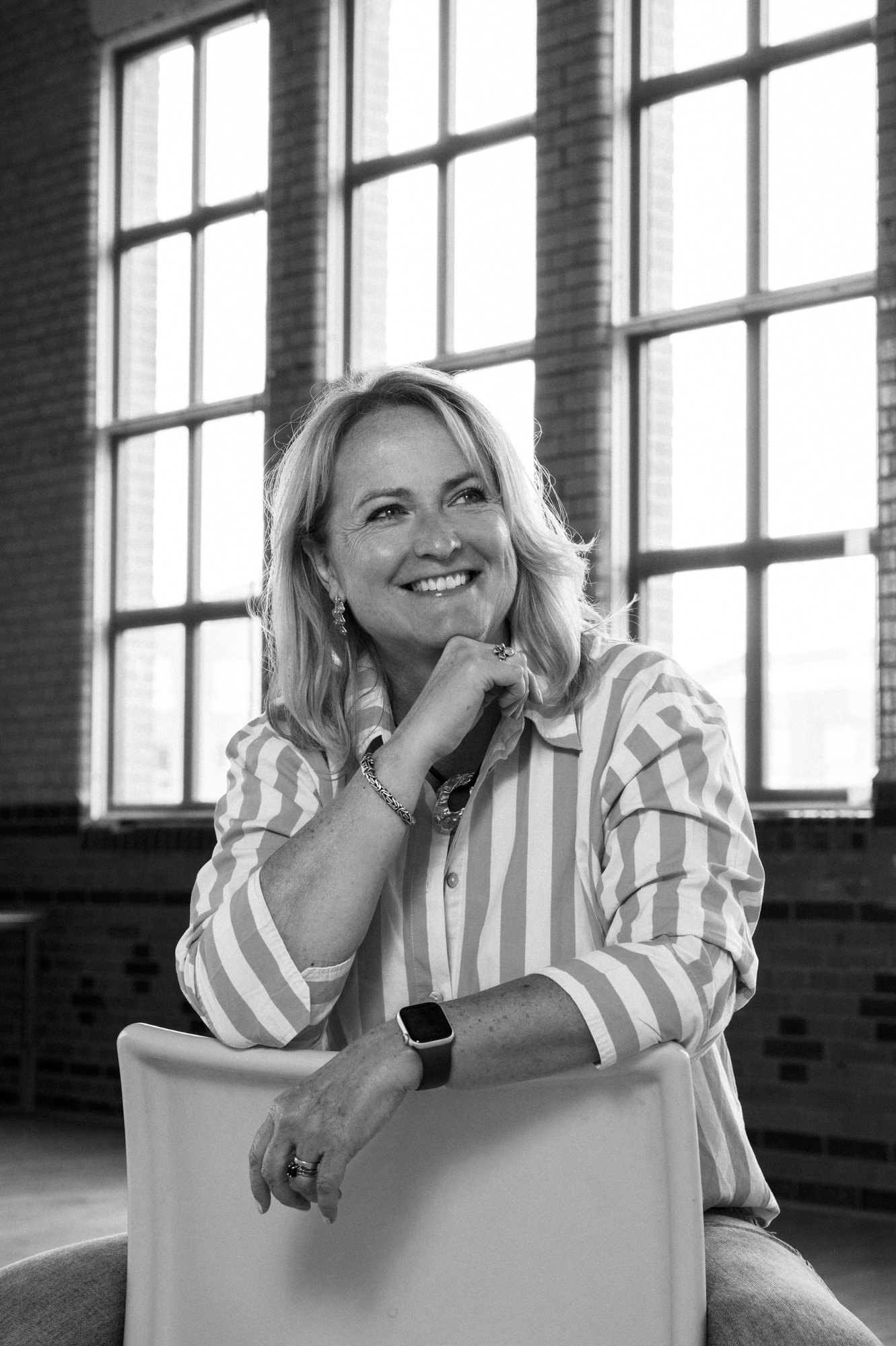 Lachende vrouw met blauwe en witte gestreepte blouse, zittend op een witte stoel binnen in een ruimte met grote ramen en bakstenen muren, zwijgzaam in zwart-wit