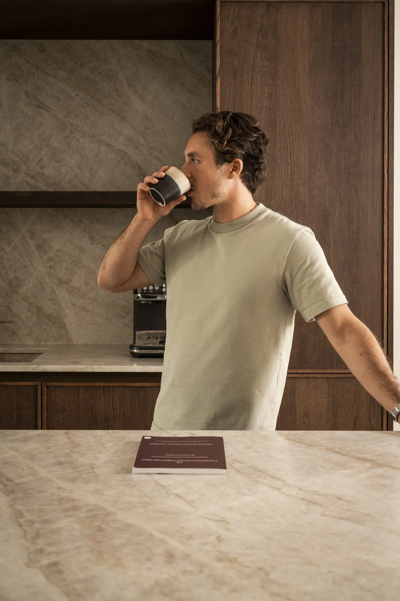 Man die koffie drinkt in een keuken met houtaccenten, een beige T-shirt en een horloge, met een boek op de keukentafel.