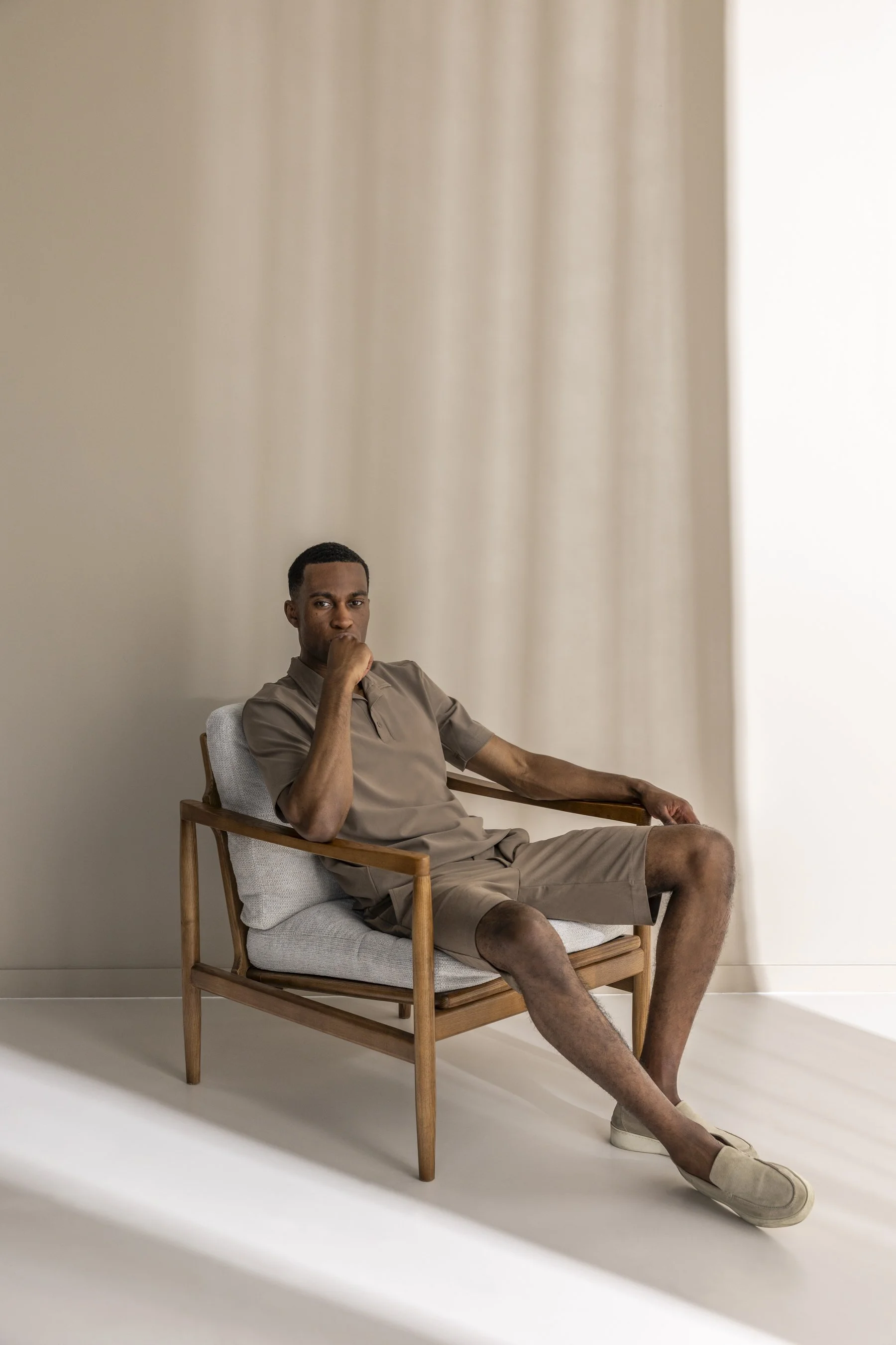 Man zit ontspannen in een houten armstoel met lichtgekleurde kussens, gekleed in een beige outfit, in een minimalistische kamer met een beige muur en witte vloer.