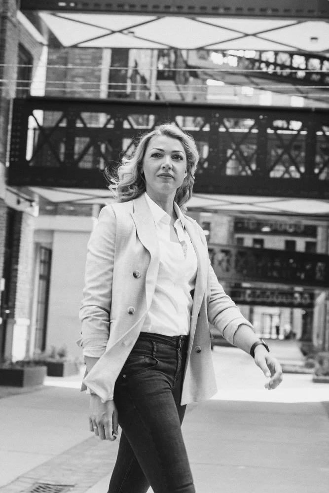 Een vrouw met blond haar loopt door een stedelijk gebied, gekleed in een witte blouse en een lichtgekleurde blazer, op een straat met gebouwen en bruggen op de achtergrond.