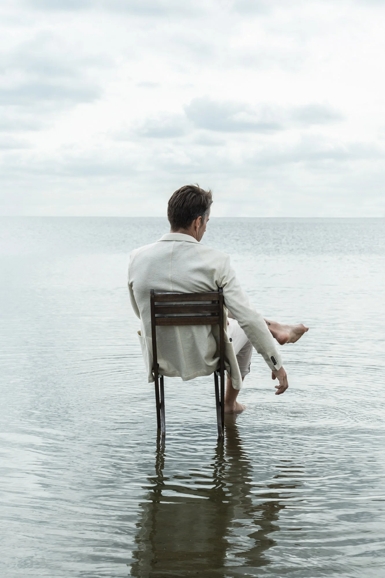 Man zittend op een houten stoel met zijn benen in het water, kijkend naar de horizon bij een meer of oceaan, bewolkte lucht