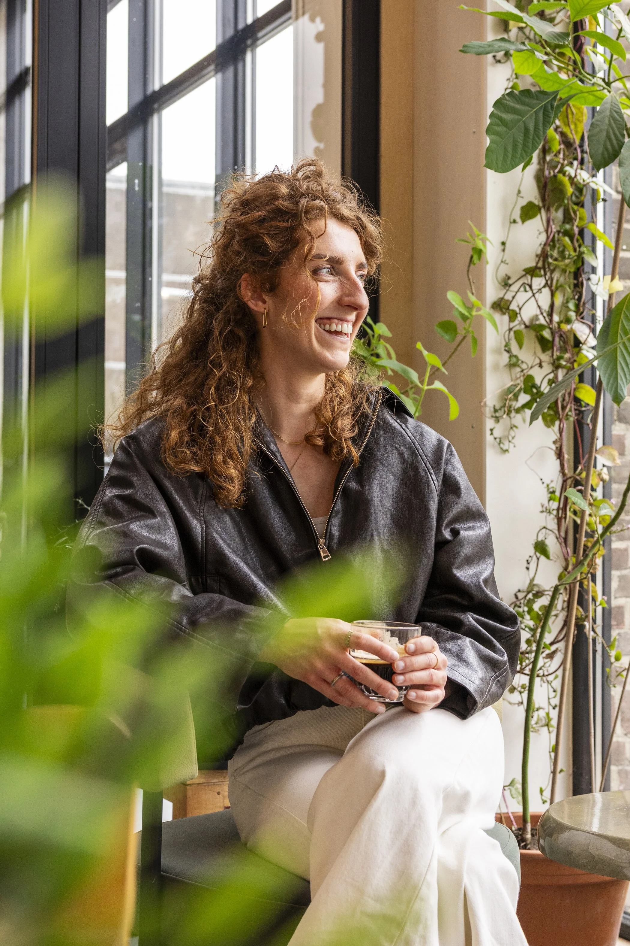 Vrouw met rood krullend haar zit binnenshuis, lachend, terwijl ze een glas met een donker drankje vasthoudt, omringd door groene planten, naast grote ramen.