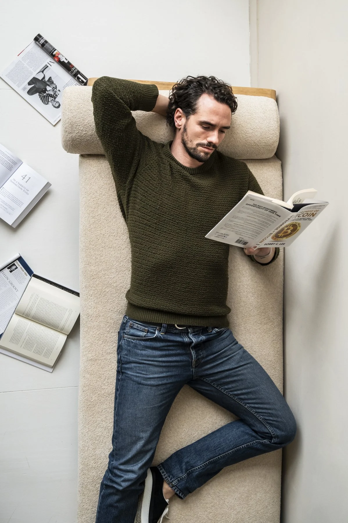Man ligt op een sofa, leest een boek met een gouden Bitcoin-logo op de omslag, met meerdere open boeken en tijdschriften rondom hem.