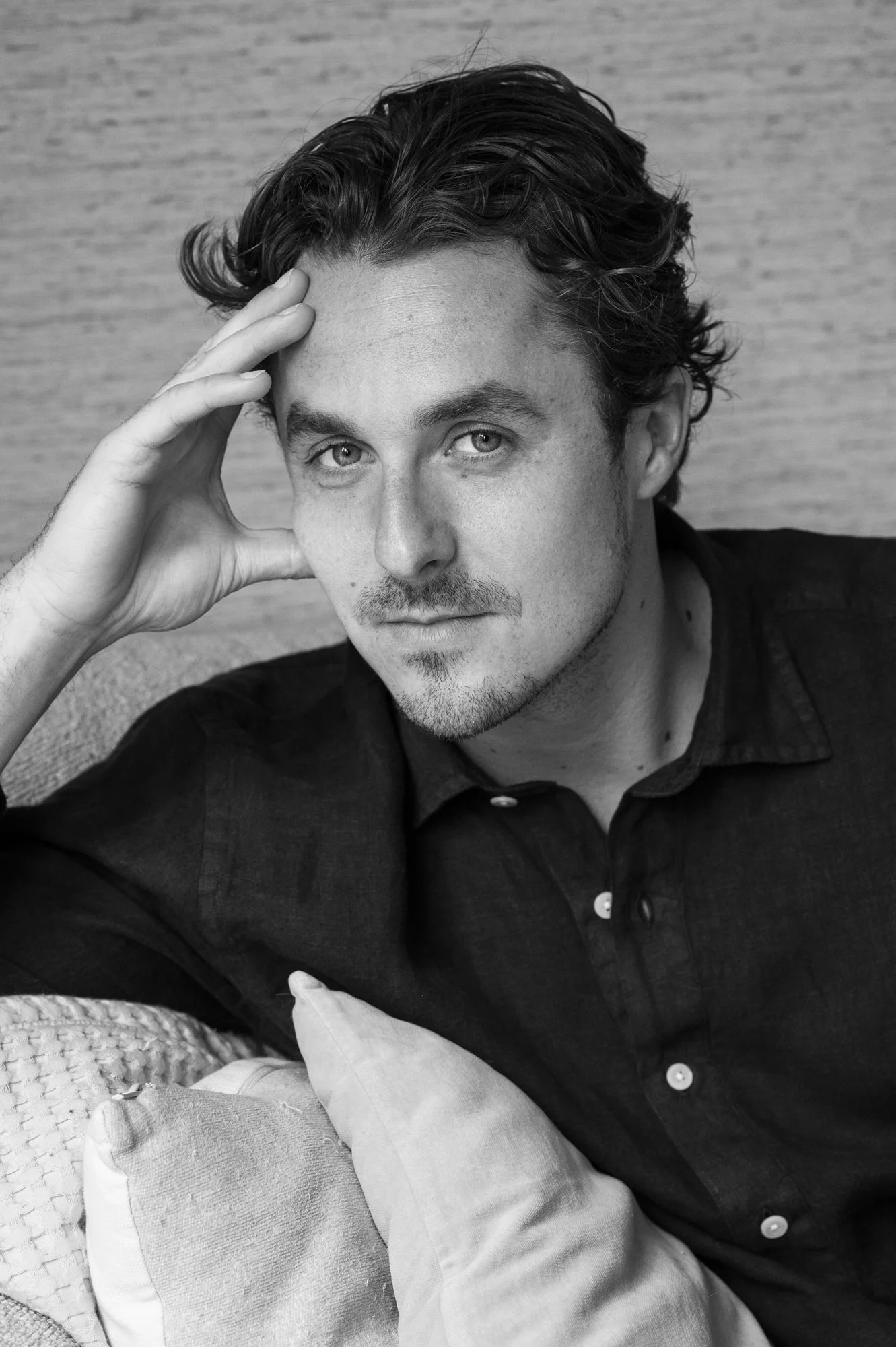 Zwart-wit foto van een man met halflang haar, lichte huid, lichte ogen en een lichte stoppelbaard, die naar de camera kijkt. Hij rust zijn hoofd op zijn hand en draagt een donker overhemd. Hij zit op een bank met kussens.