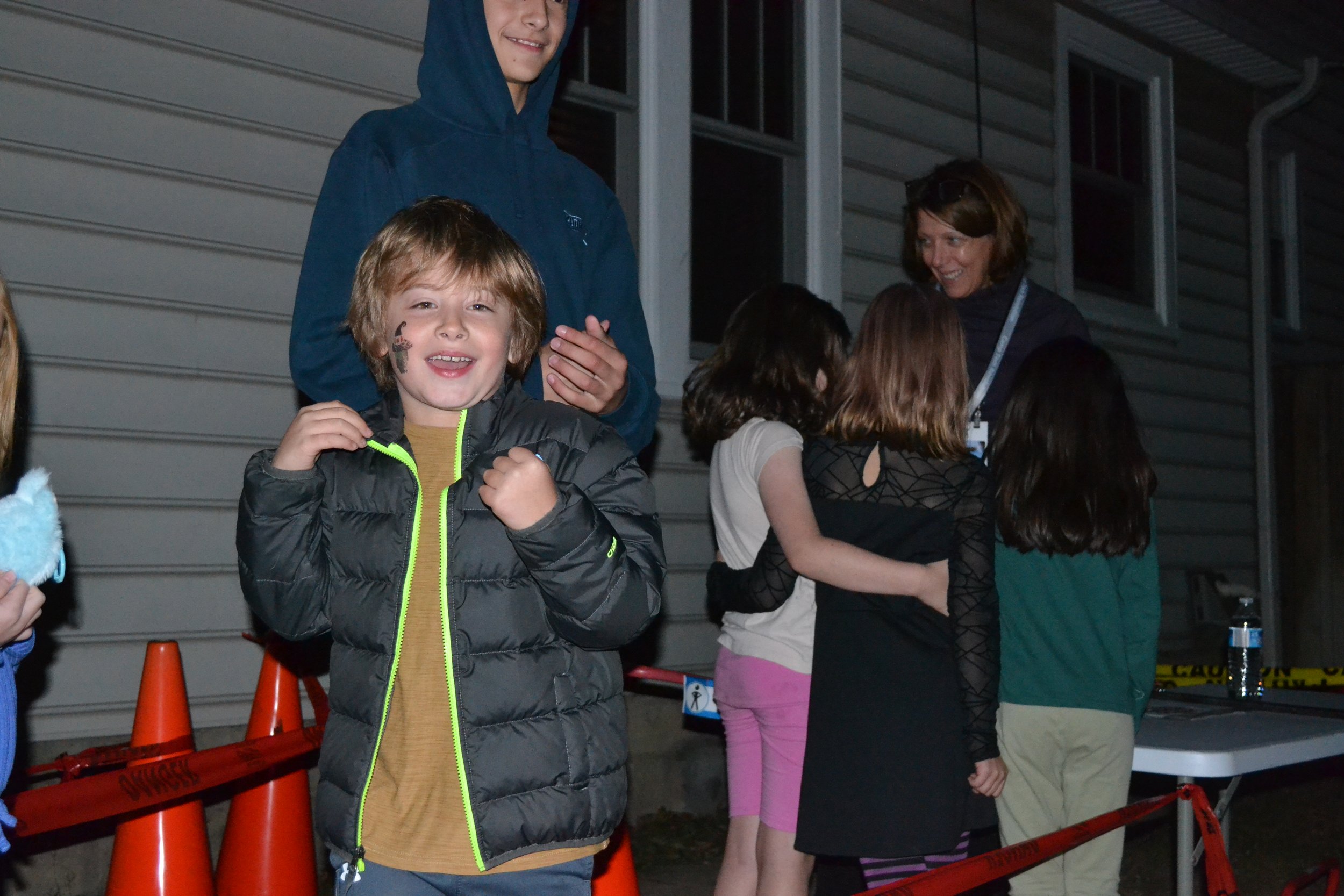 A group of children and two adults gather outside at night near orange cones and caution tape, with one young boy smiling and showing a fist as he stands in front.