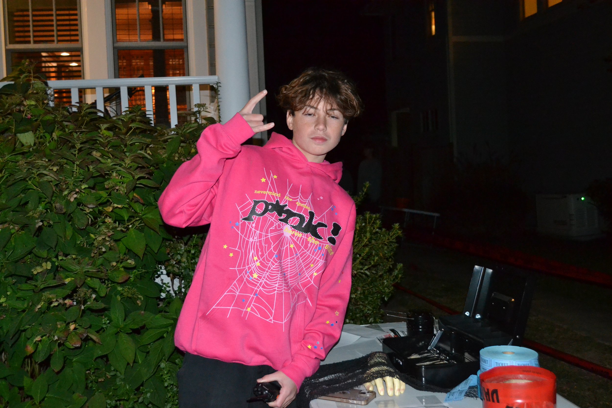 A young person with short, curly hair making a playful peace sign gesture near the head with a pink hoodie that has a spider web graphic and the word 'porg!'. They are standing outdoors near some bushes, with a table and electronic equipment on it, late evening or night setting.