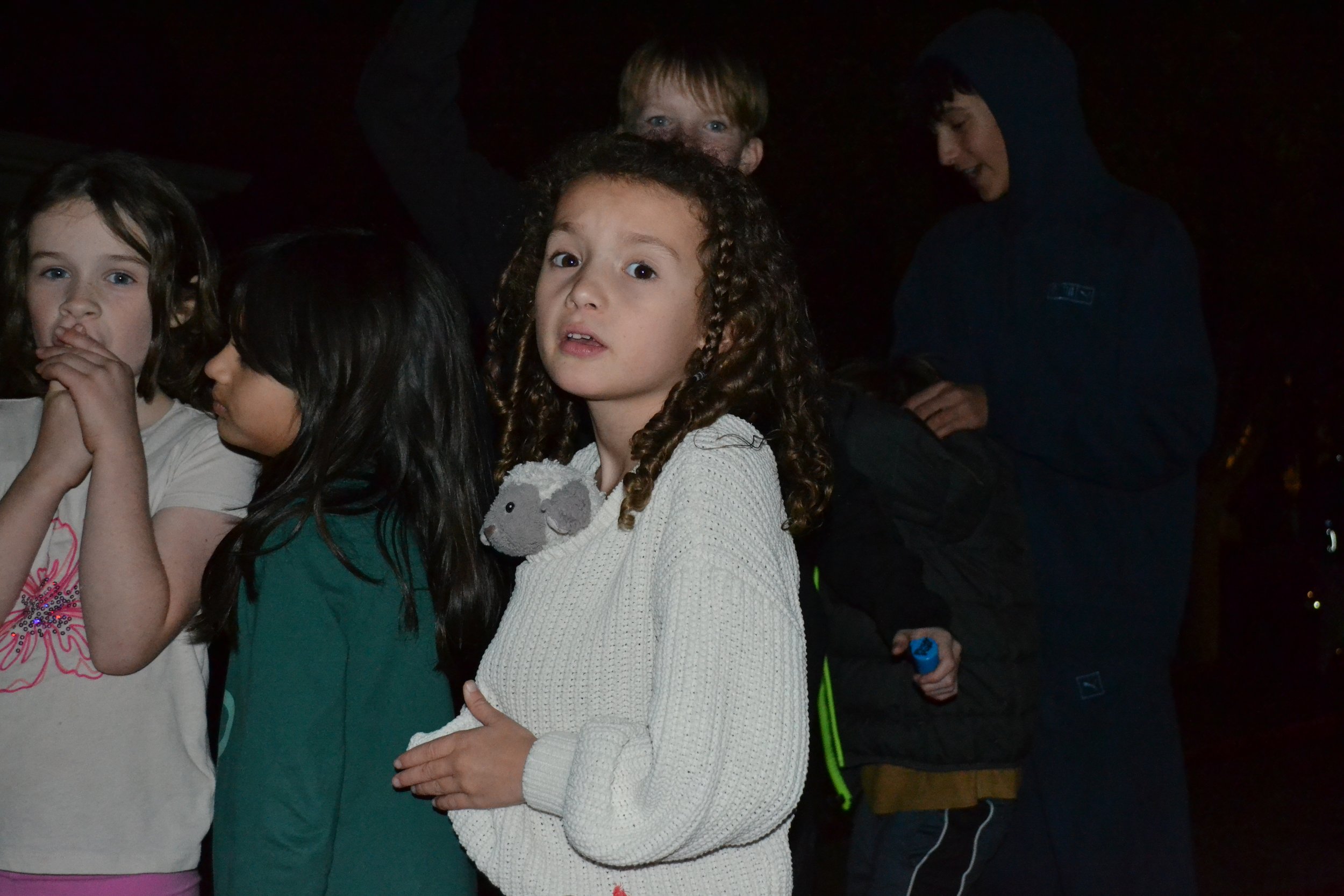 Group of children gathered outdoors at night, with one girl in the center holding a stuffed animal, looking at the camera, and others around her engaged in conversation.