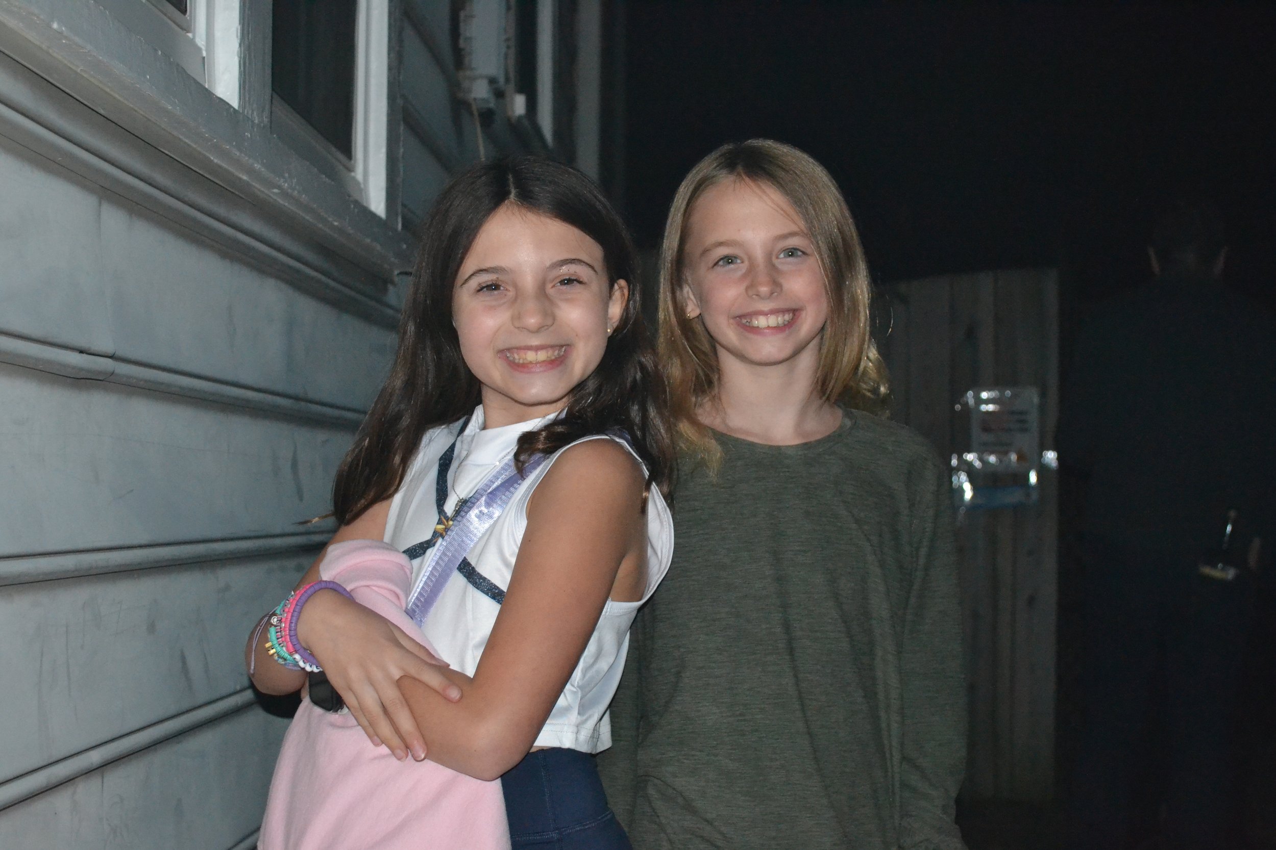 Two young girls smiling and standing outside at night, with a garage door and a wooden fence in the background.