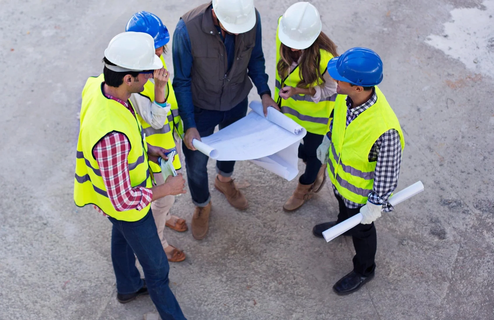 A group of engineers and construction workers wearing safety helmets and reflective vests gathered around a set of blueprints at a construction site.