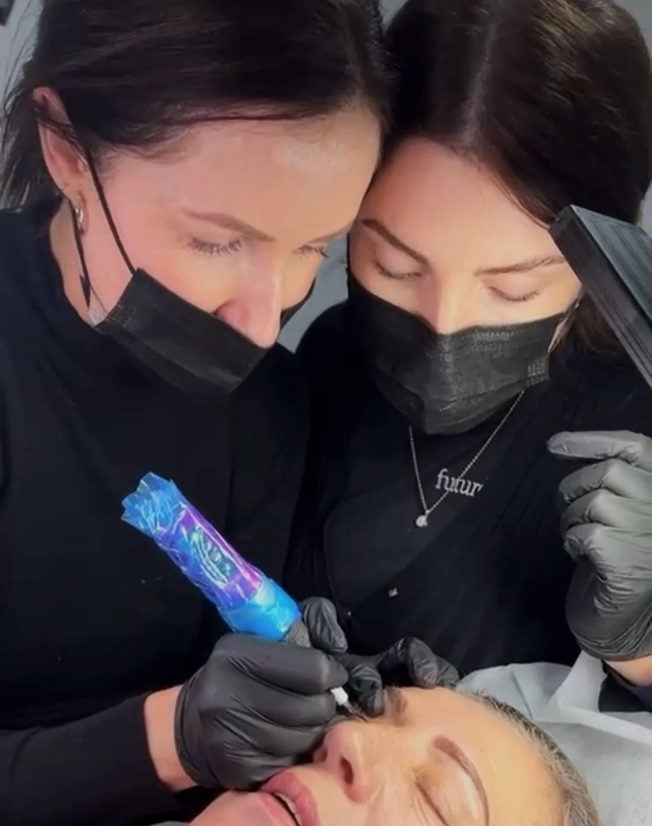 Two women wearing black masks and gloves are performing a cosmetic injection on a woman's lips. They are holding a syringe with a colorful needle design and are focused on her face.