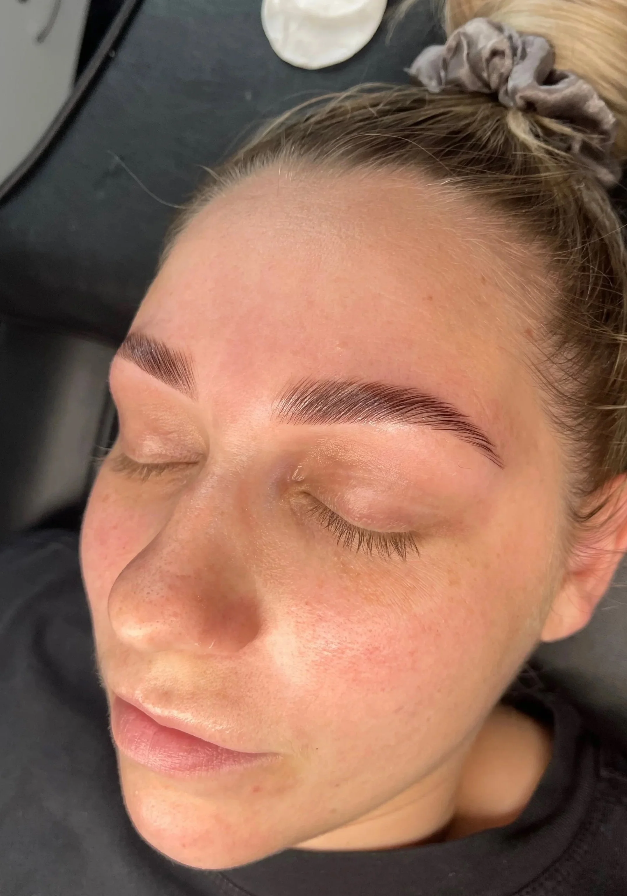 Close-up of a woman's face with freshly done eyebrow microblading, eyes closed, lying down with hair in a bun, on a black surface.