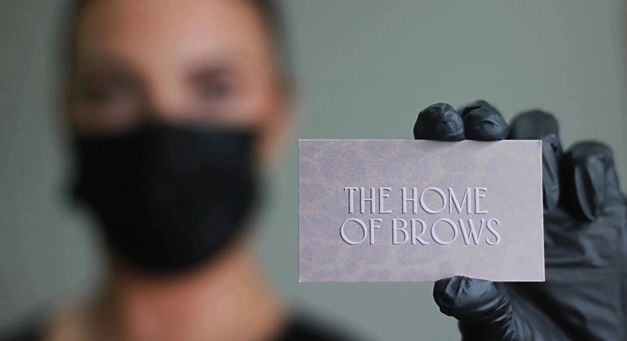 A person wearing a black mask and black gloves holds a card that reads "The Home of Brows." Their face is out of focus, with only the eyes partially visible.