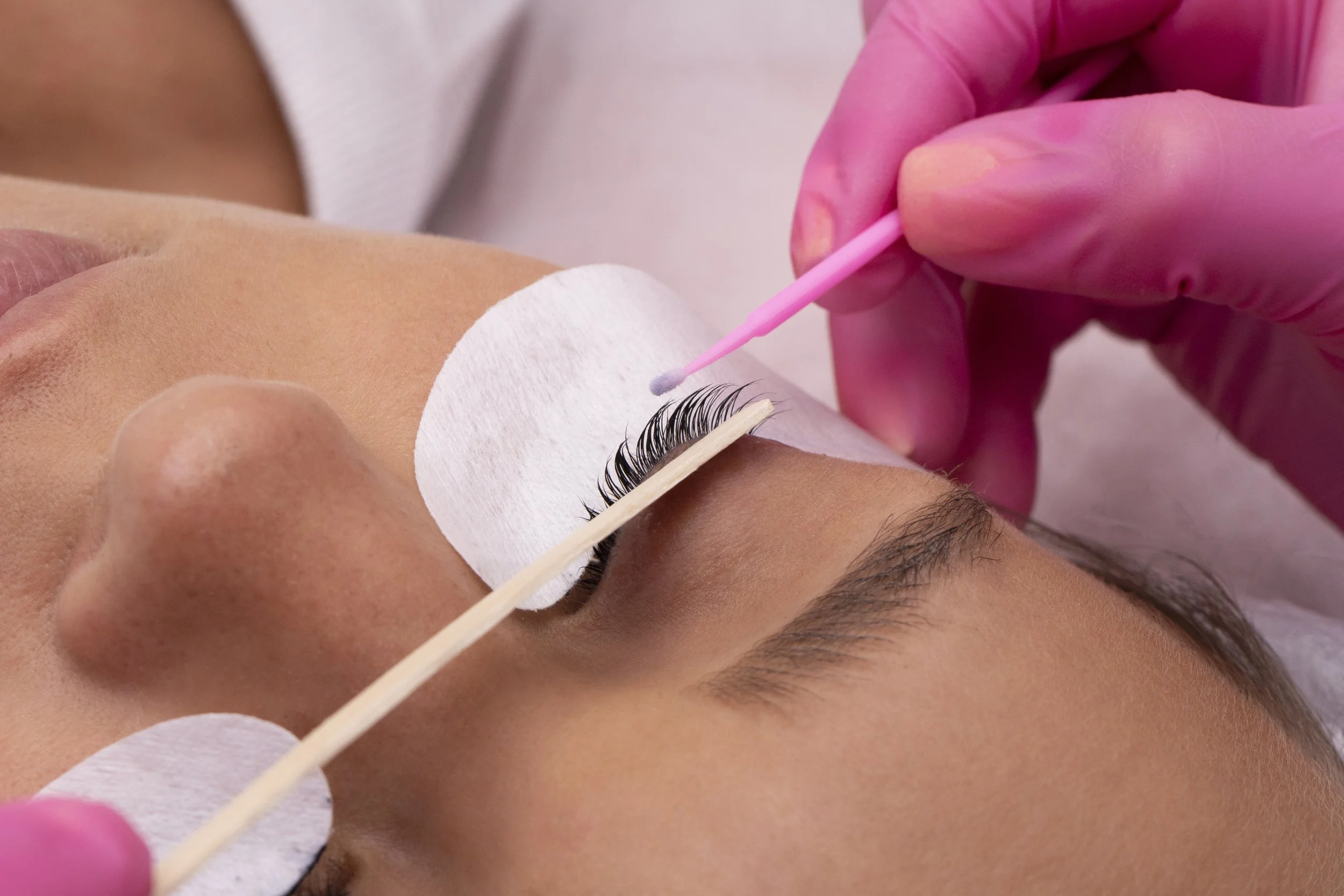 Close-up of a person receiving eyelash extension application, with black synthetic eyelashes, cotton pads on the skin, and a pink-tipped applicator tool held by gloved hand.