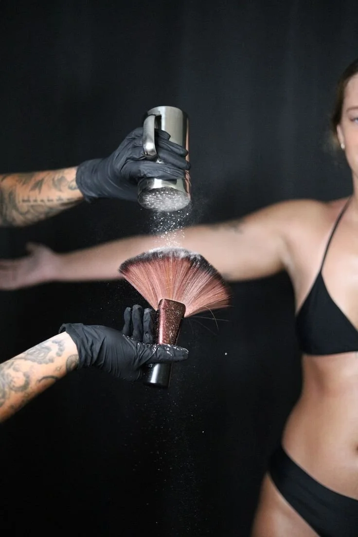 Someone sprinkles powder onto a makeup brush held by another person in black gloves, with a woman in black lingerie in the background.