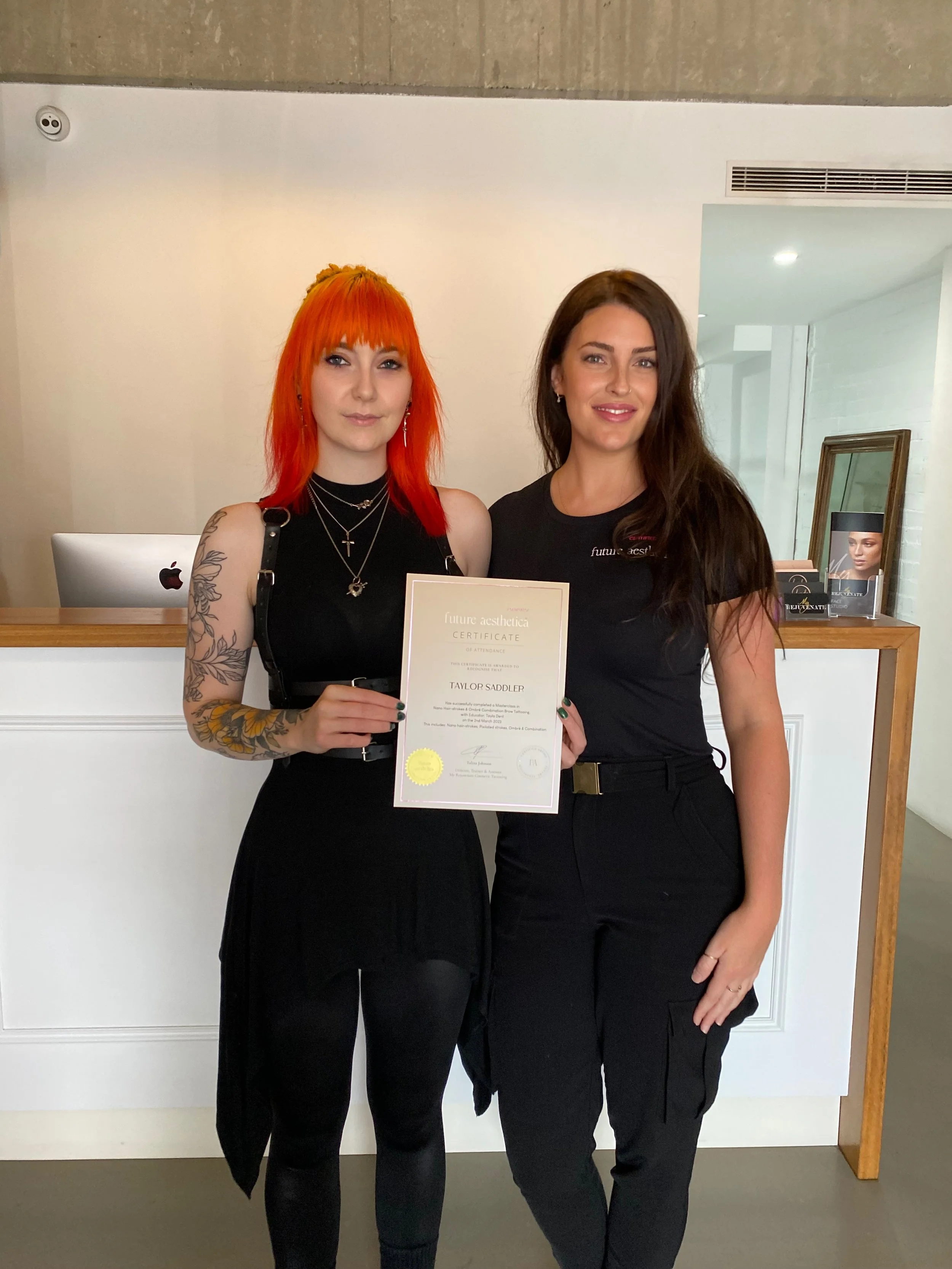 Two women standing in front of a reception desk, one holding a certificate, smiling for the camera. The woman on the left has bright orange hair and tattoos on her arm, wearing black clothing. The woman on the right has long dark hair, also dressed in black. The background includes a framed photo on a stand and a computer on the reception desk.