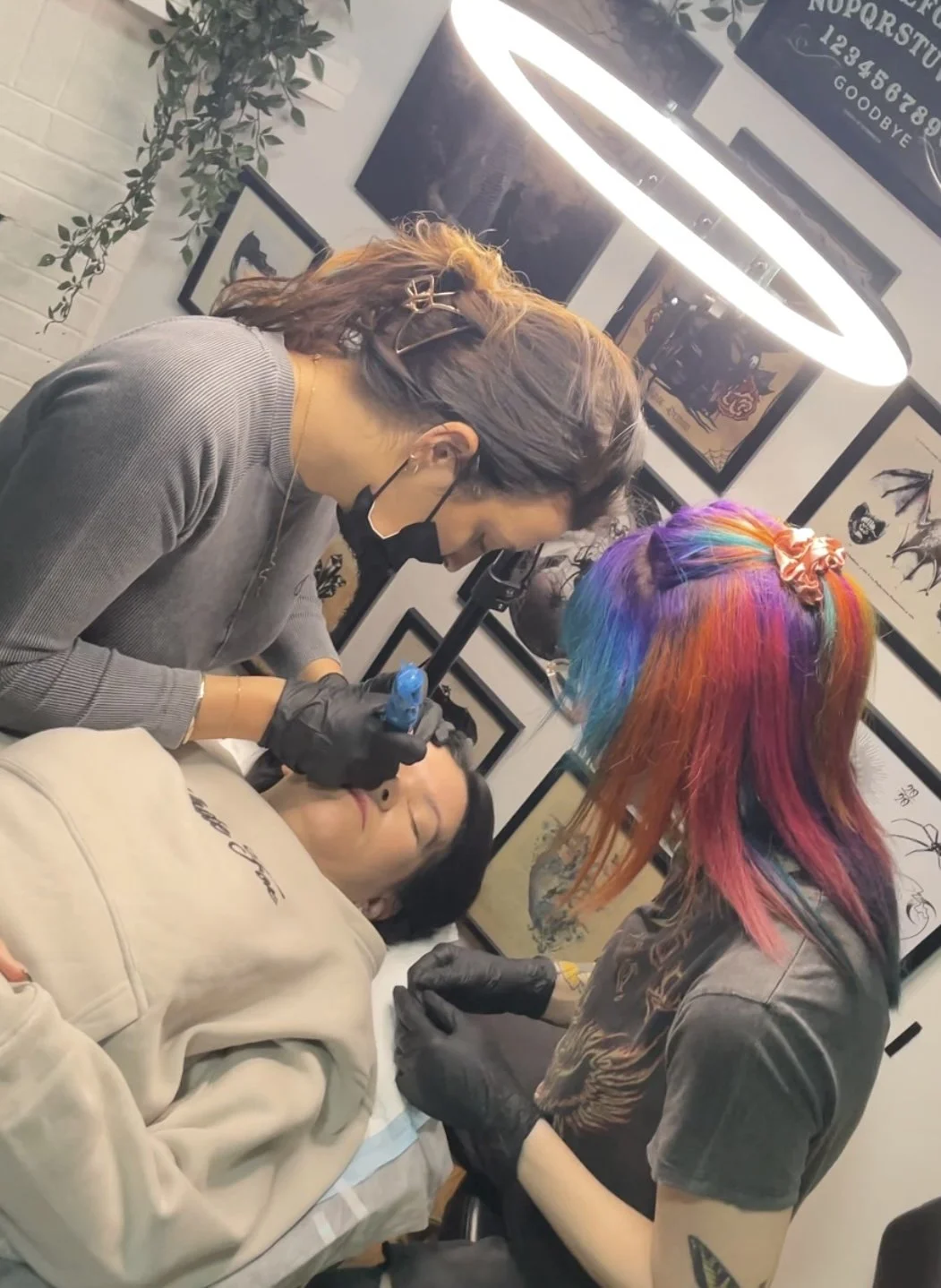 A woman with rainbow-colored hair receiving a tattoo from a tattoo artist in a studio with framed artwork on the walls.