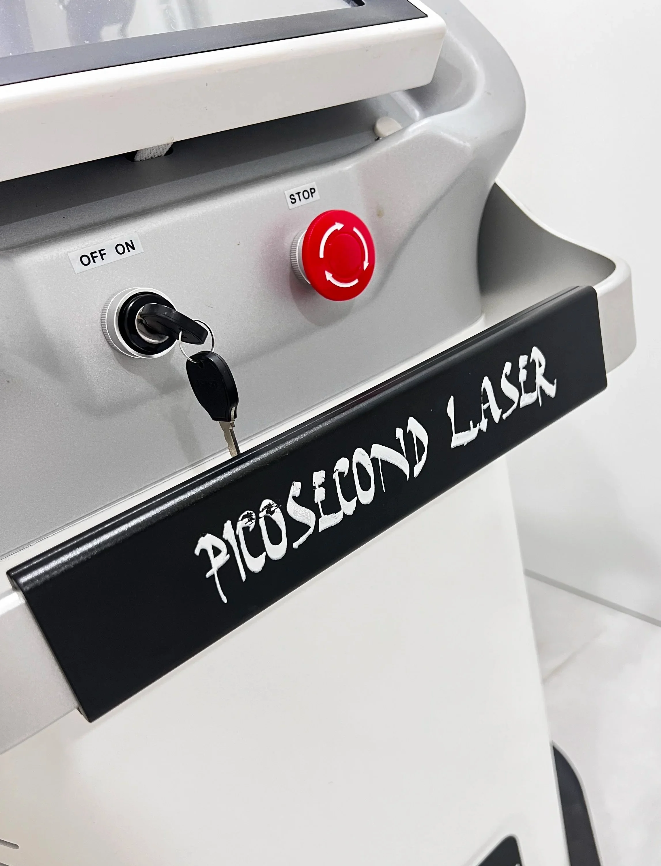 Close-up of a PicoSecond Laser machine control panel with an ON/OFF switch, a red emergency stop button, and a black sign with white text reading "PICOSECOND LASER".