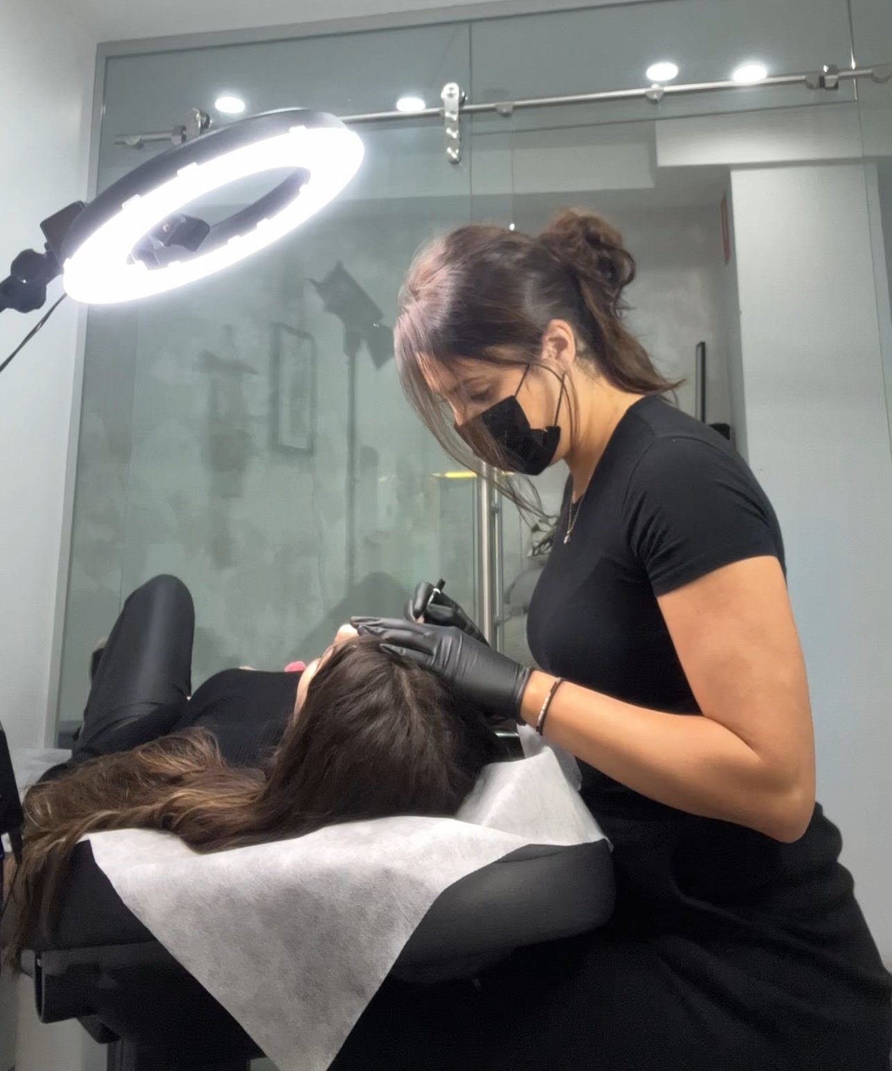 A woman with dark hair, wearing a black mask, black gloves, and black t-shirt, is performing a tattoo procedure on a client lying on a tattoo bed.