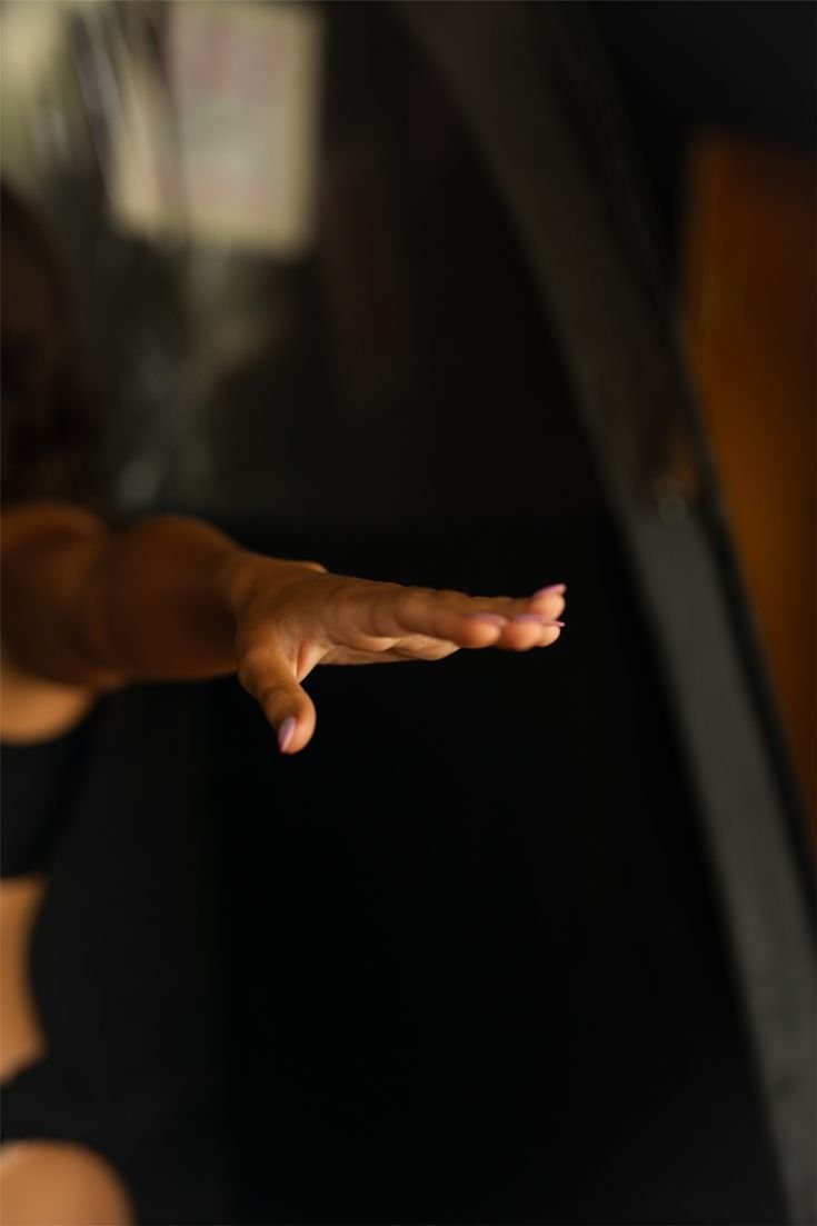 A person's hand gracefully stretched out above a black piano keyboard, with a blurred background of a room with a window.