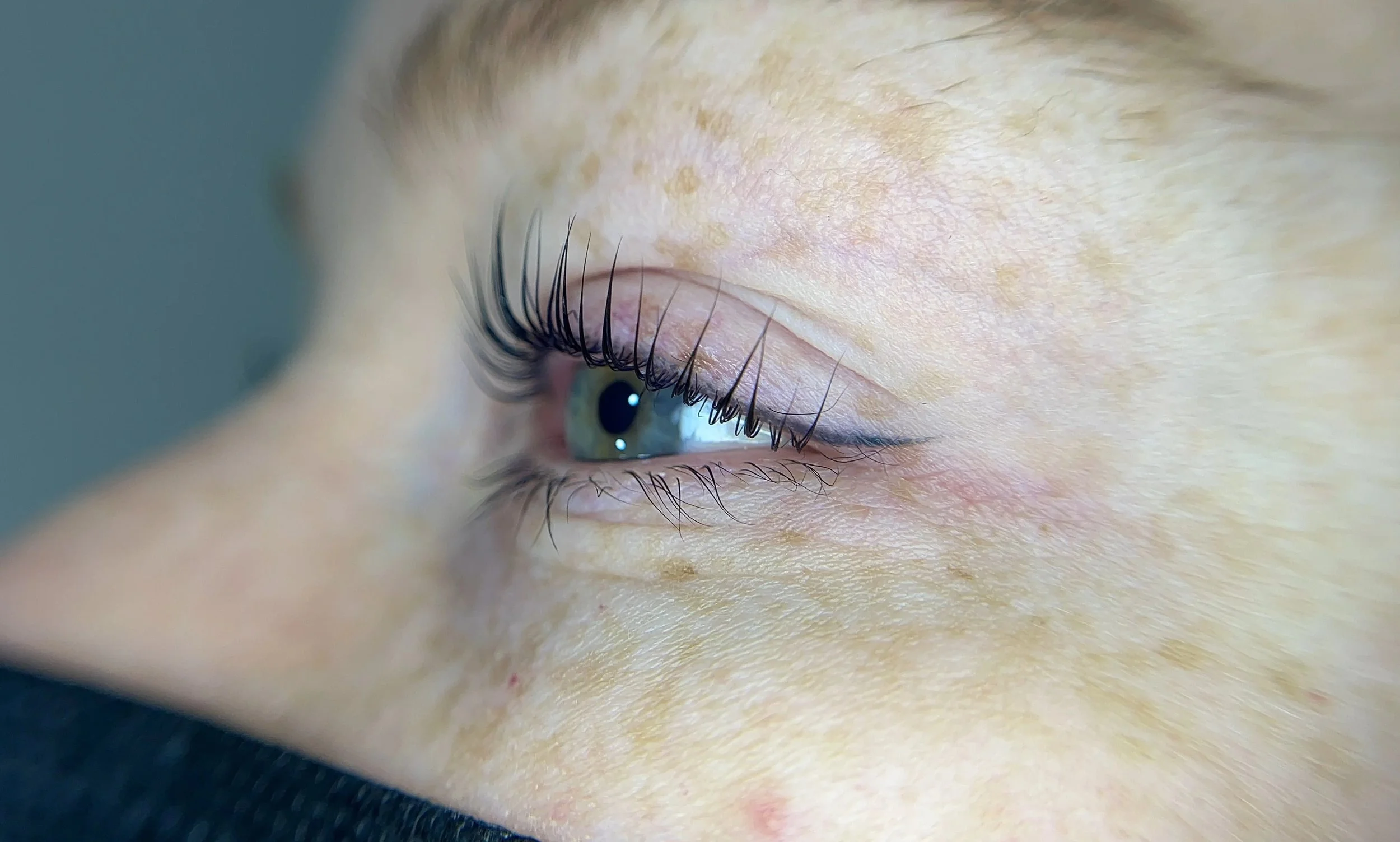 Close-up of a person's eye with long curled eyelashes, light blue iris, and skin showing freckles and fine details.