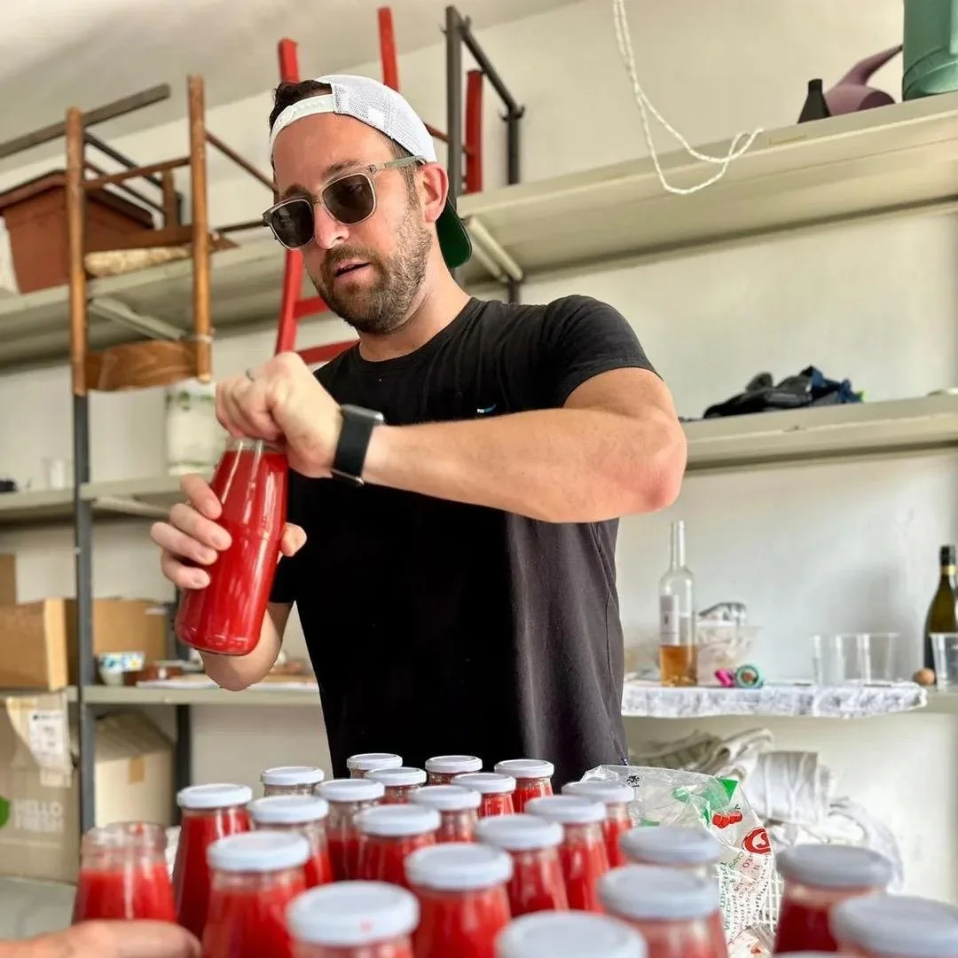A man wearing sunglasses, a black t-shirt, and a backwards white cap is closing a red bottle of passata among many similar bottles on a table. He is in a garage or storage room with shelves holding various items behind him.