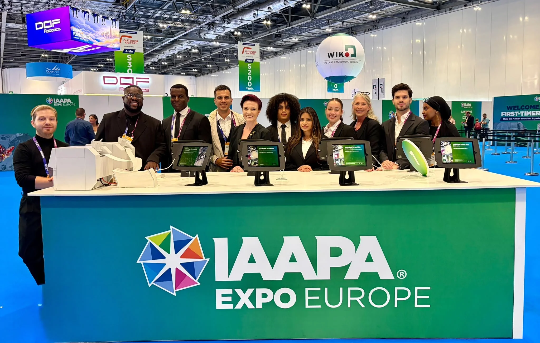 Group of people standing behind a booth with a green sign that reads "IAAAPA Expo Europe" displaying electronic devices at a trade show.