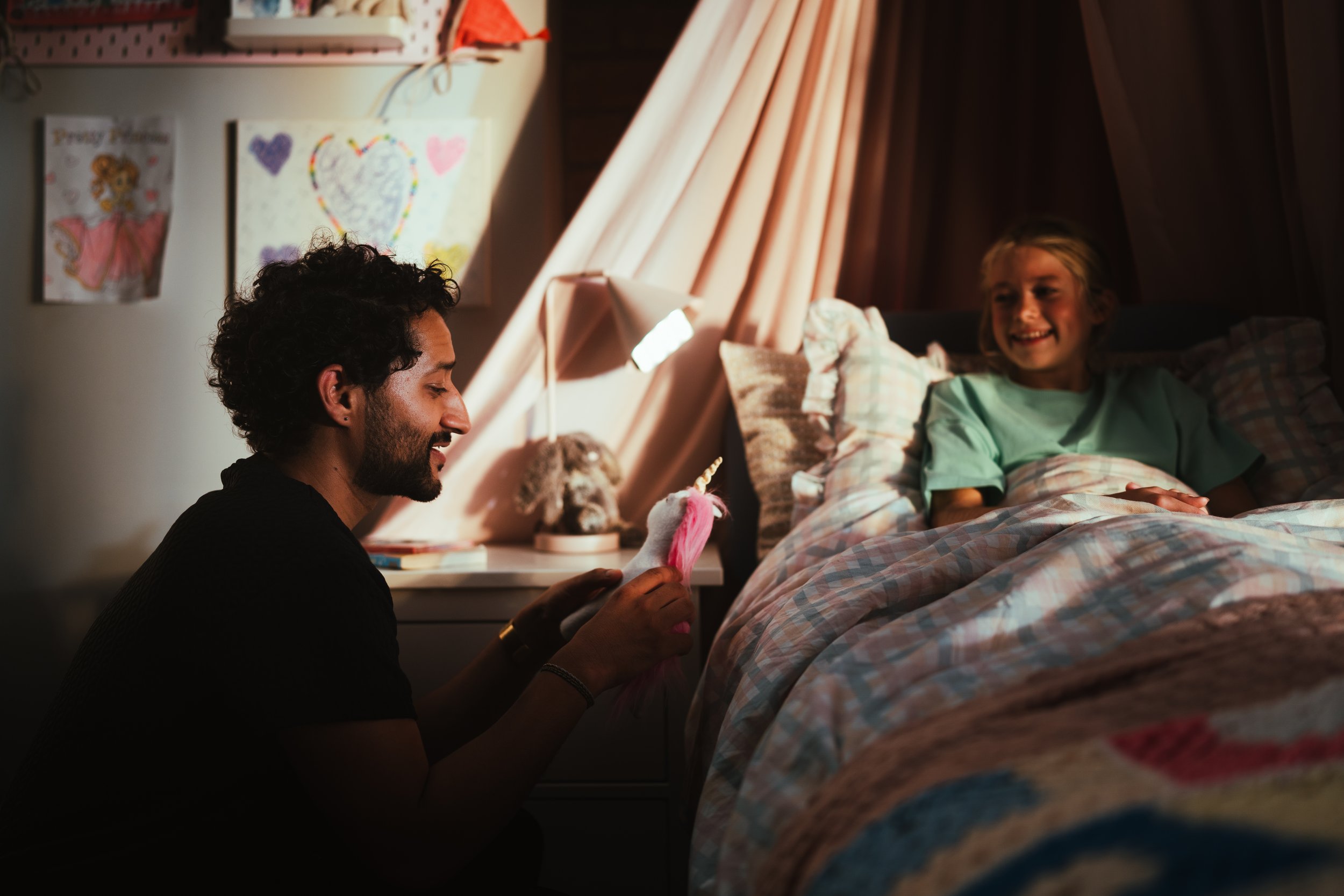 A man giving a unicorn plush toy to a girl in bed, smiling with a heartwarming moment in a cozy bedroom.