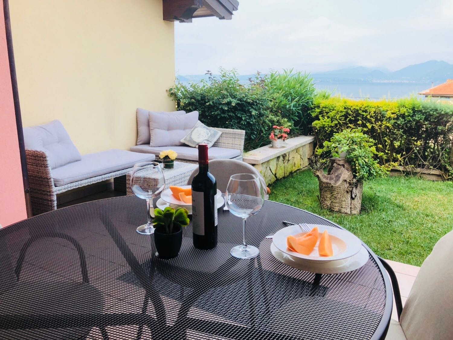 An outdoor patio scene with a round table set for wine and cheese, a basket of grapes, and a wine bottle. In the background, there is a cushioned sofa, green plants, and a scenic view of mountains and water.