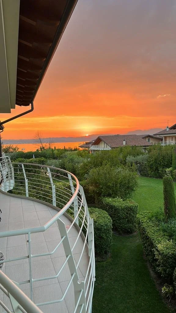View from a balcony overlooking a green backyard at sunset with orange and pink sky, houses in the distance, and mountains.