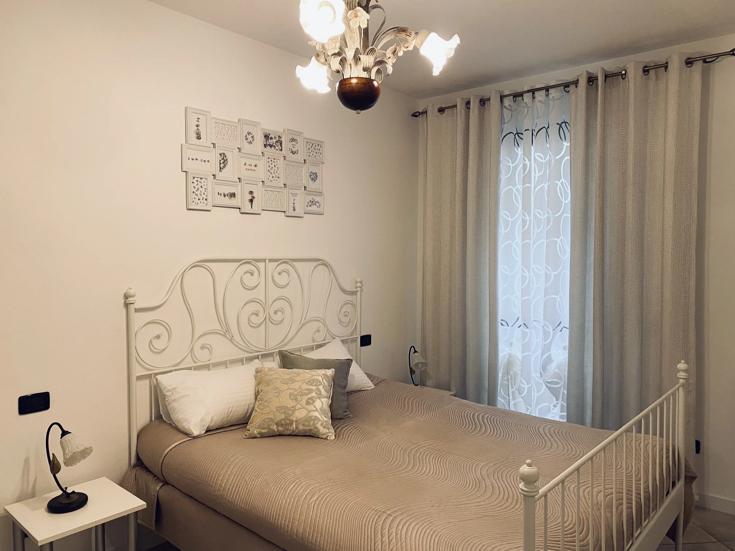 A bedroom with a white metal bed frame, beige bedding, and pillows. There is a small white nightstand with a black lamp on the left side, white walls, a window with sheer and beige curtains, and a framed collage of small picture frames above the bed.