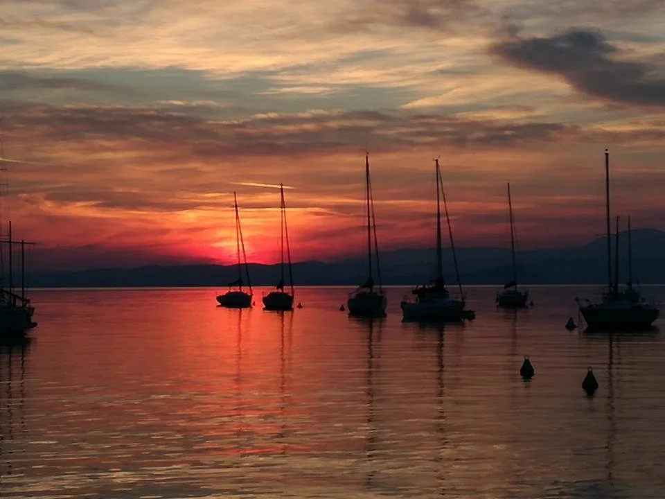 A sunset over a calm body of water with sailboats anchored, and mountains in the distance.