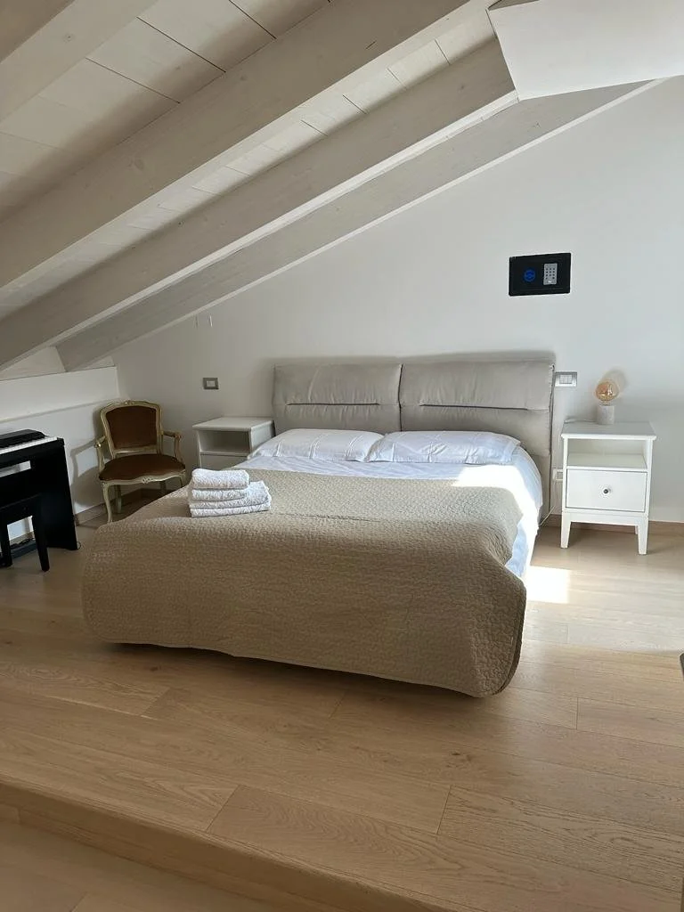 A neatly made bed with white pillows and beige quilt, topped with folded white towels, in a bright bedroom with a sloped ceiling, nightstands on both sides, a small white chair, a black piano, and a safe on the wall.