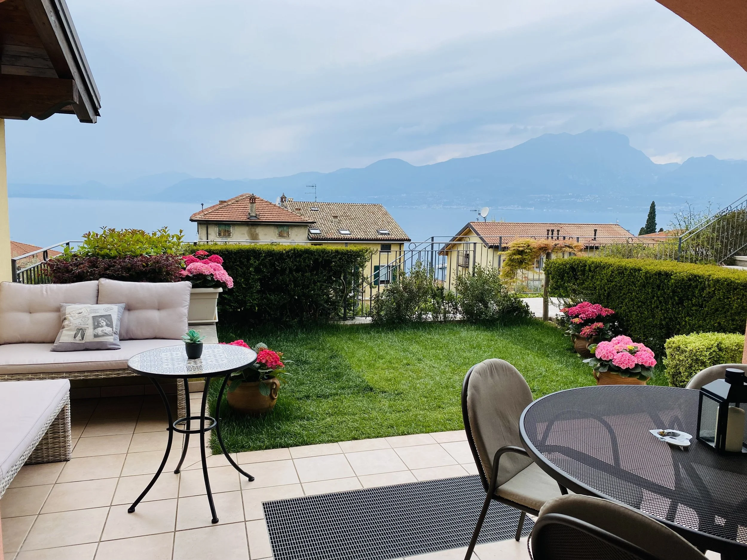 Terrace with outdoor furniture, including a round table, chairs, and a cushioned sofa. Potted pink flowers and greenery decorate the space, overlooking a view of houses, a lake, and mountains in the distance.