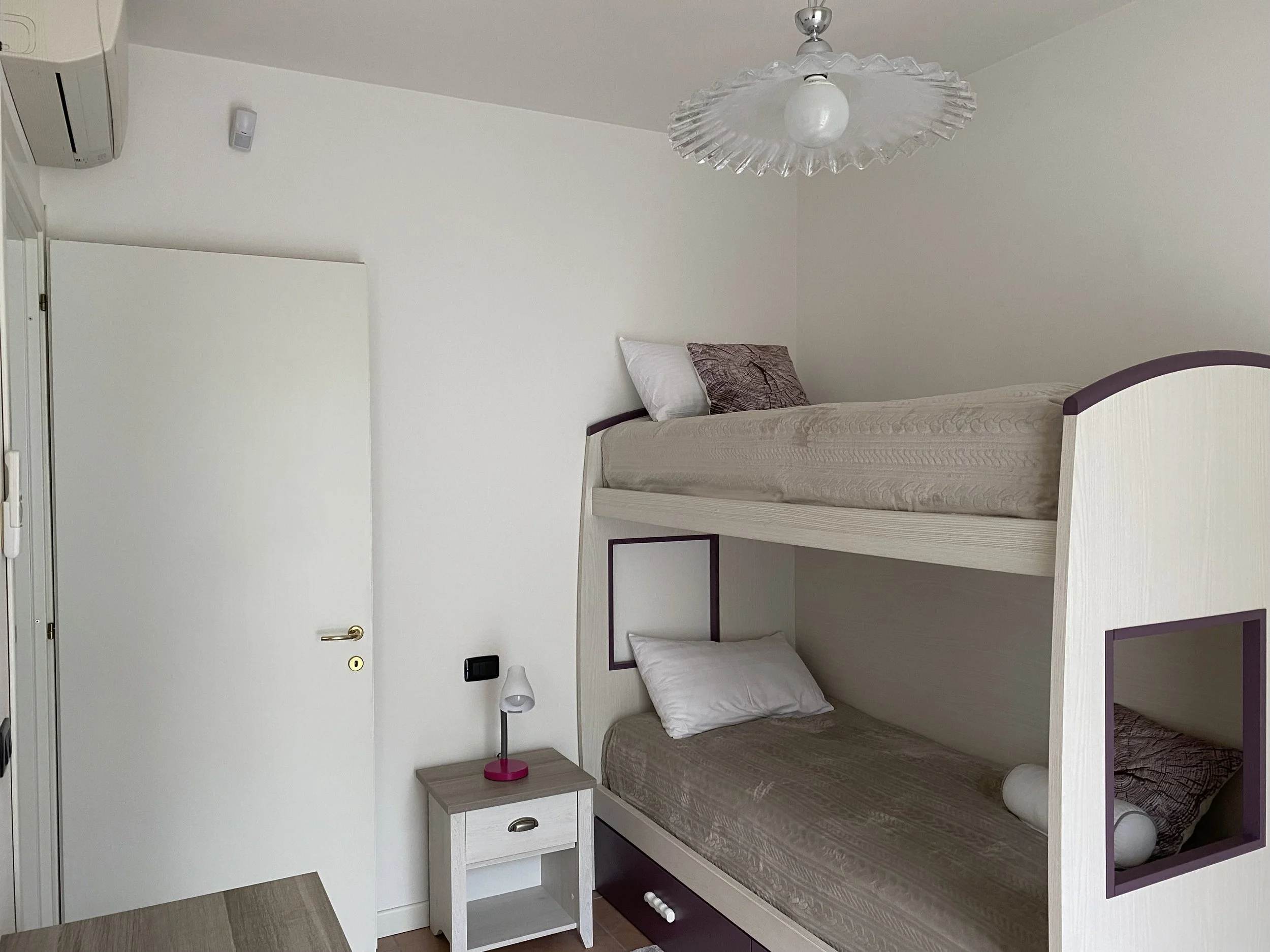 A room featuring a white bunk bed with purple accents, a small bedside table with a white lamp, and a ceiling light with a decorative glass shade. There is also an air conditioning unit on the wall and a white door.