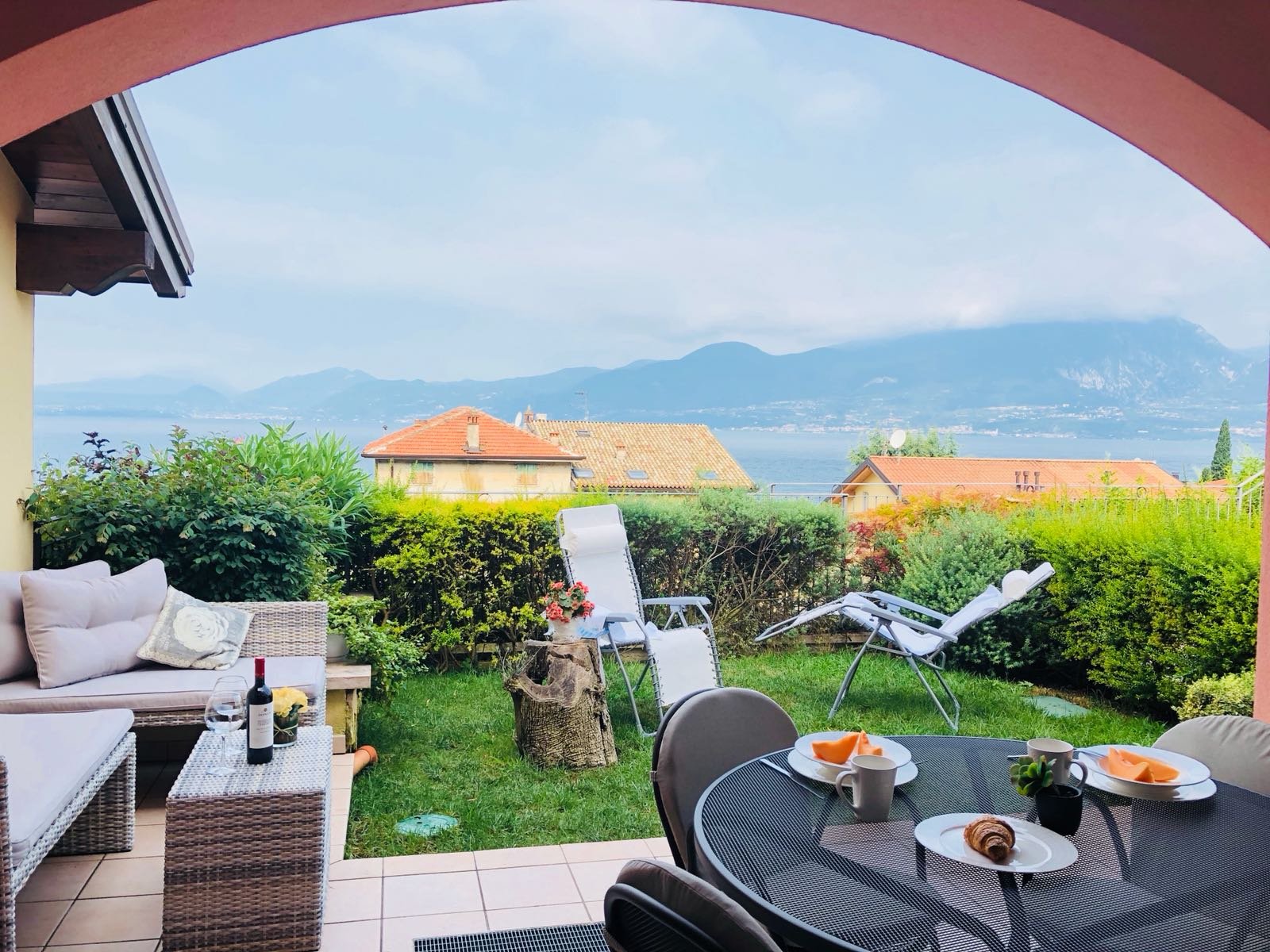 Outdoor patio with a dining table set for breakfast, surrounded by greenery with a lake and mountains in the background.