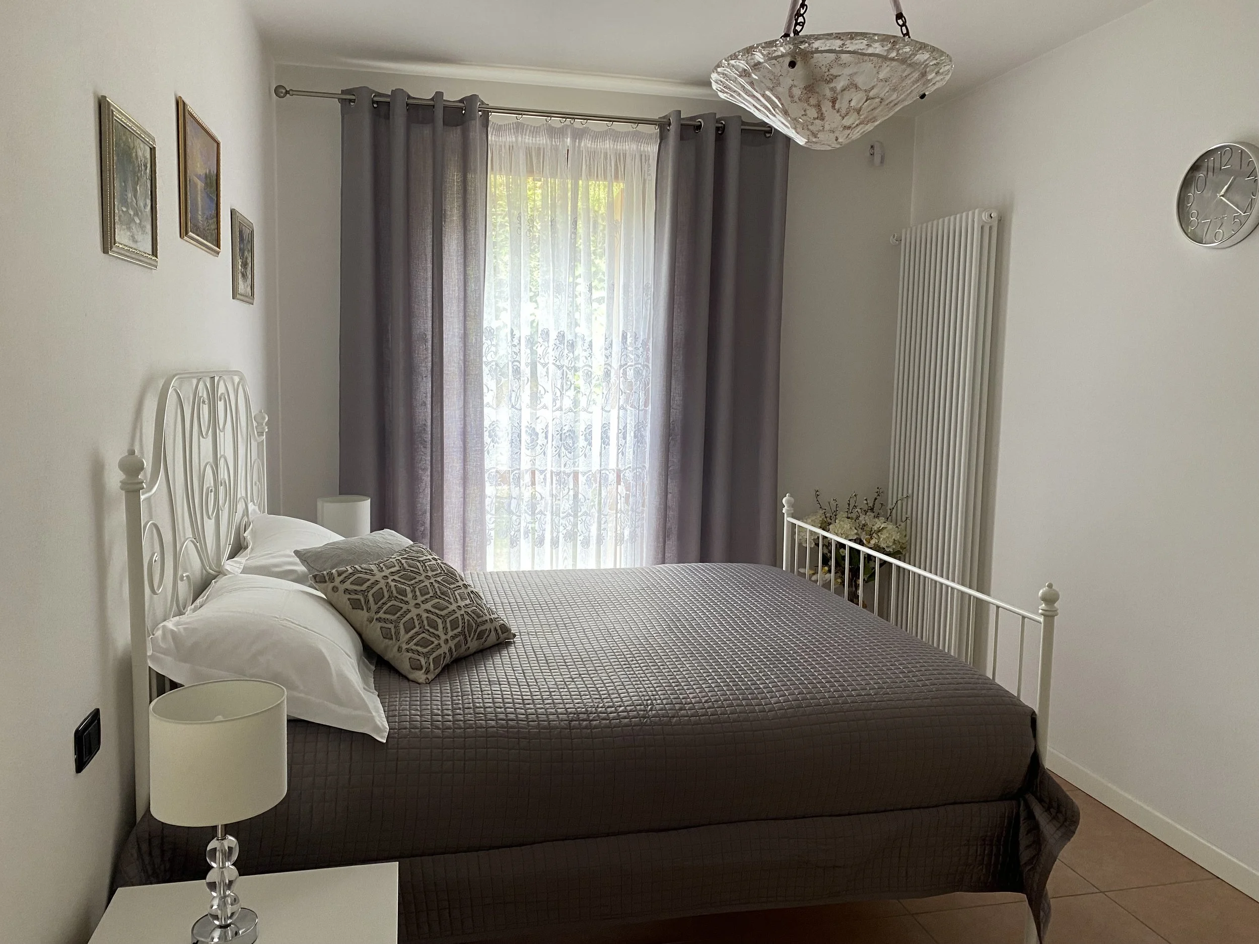 A bedroom with white walls, a white metal bed frame, brown quilted bedspread, two white pillows, an accent pillow with a geometric pattern, a bedside table with a white lamp, a window with white sheer curtains and gray drapes, a ceiling light fixture