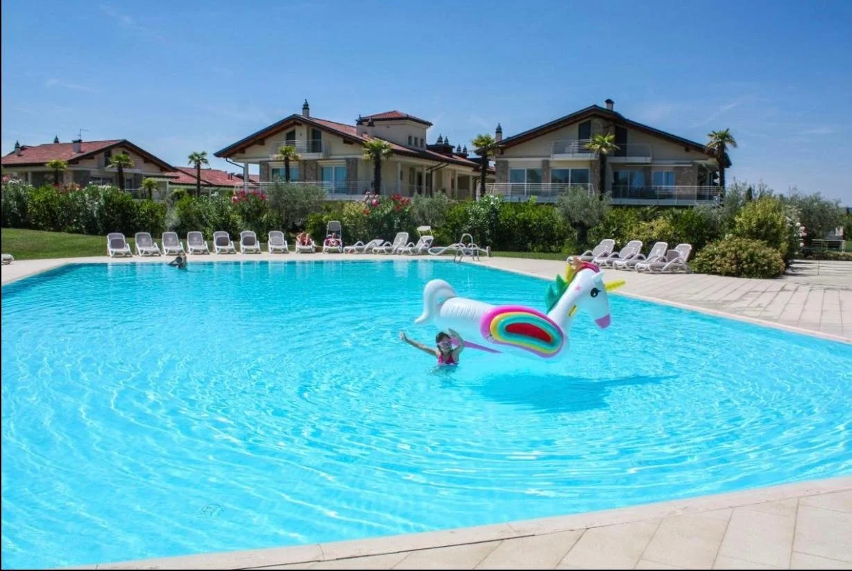 Children swimming in a large outdoor pool with a unicorn-shaped inflatable float amid lounge chairs and houses in the background on a sunny day.